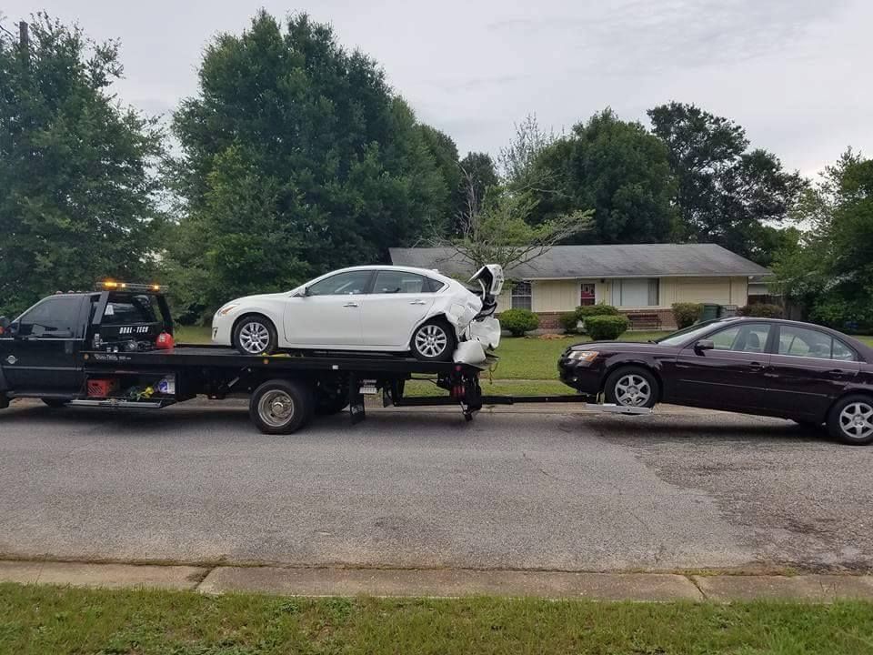 Tow truck hauling a white car after a crash, with two damaged cars on a suburban street.