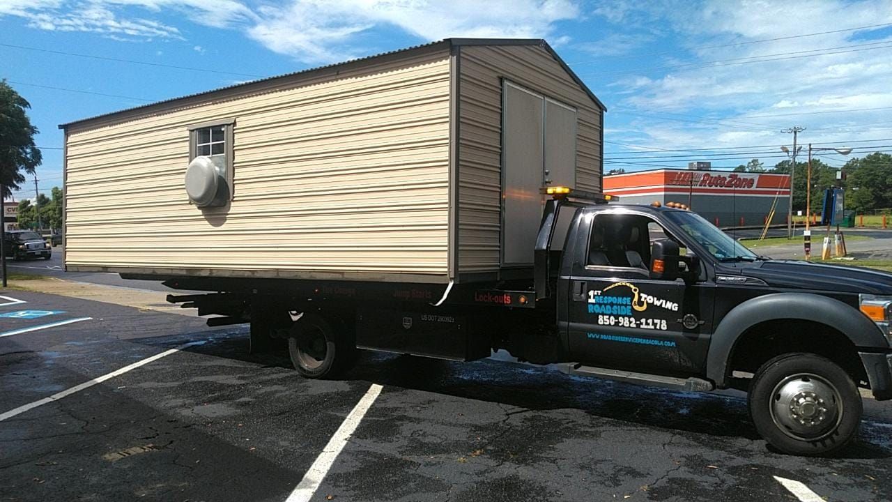 Tow truck carrying a tan mobile home on a sunny parking lot, with blue sky and painted parking lines.