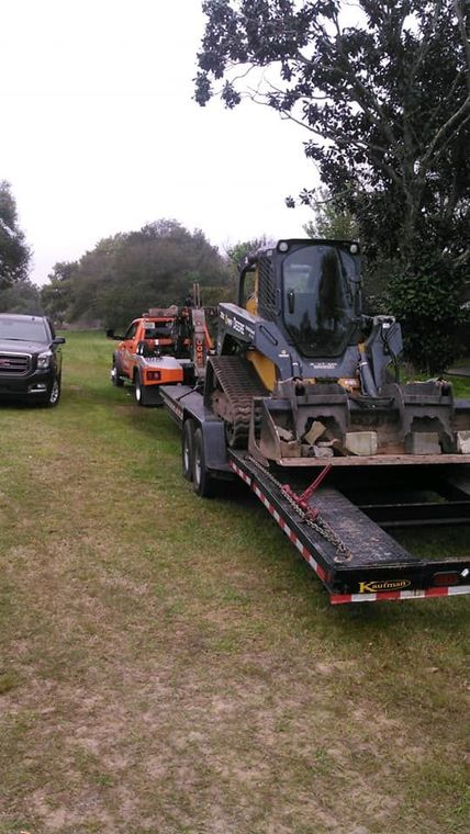 Flatbed tow truck carrying a yellow skid steer on a grassy roadside with cars lined behind it