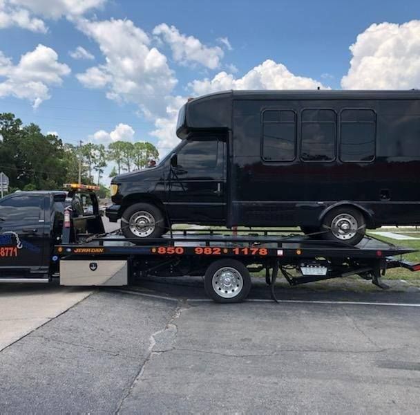 Black van loaded on a flatbed tow truck in a parking lot under a blue sky.