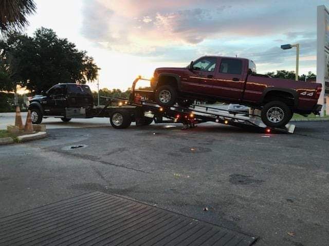 Tow truck carrying a maroon pickup beside a black SUV at sunset