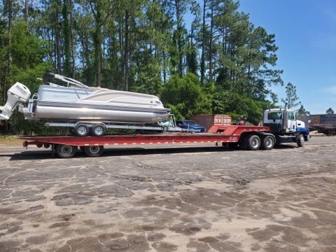 White boat on a red flatbed trailer with a blue semi truck, parked on a gravel lot by trees.
