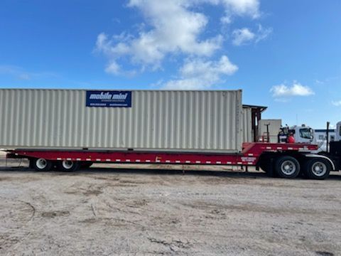 Red flatbed truck hauling a white shipping container on a gravel lot under a blue sky