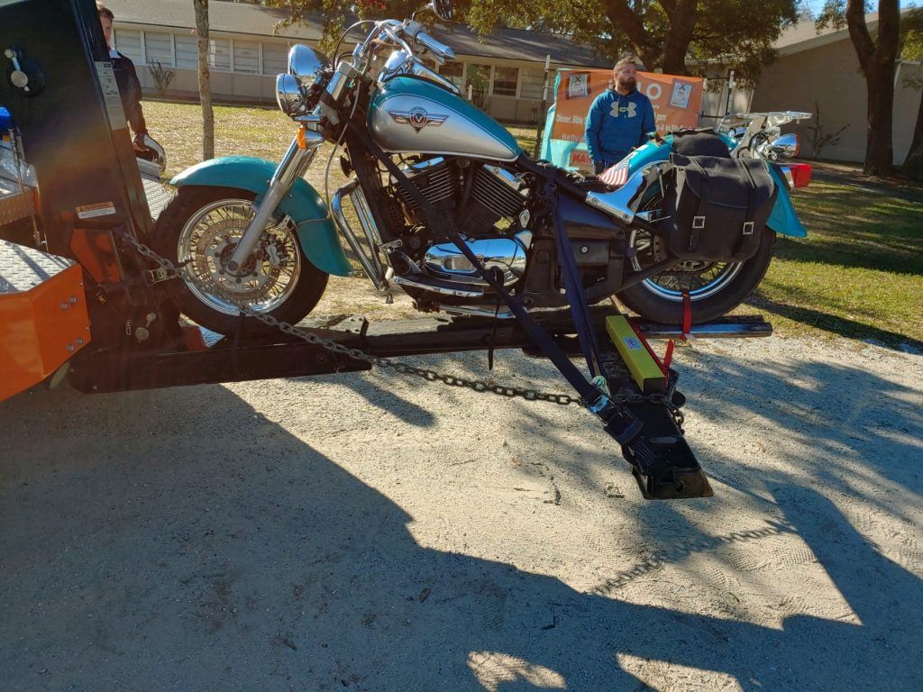 Blue custom motorcycle on a ramp outdoors, with a person standing beside it in a sunny yard.
