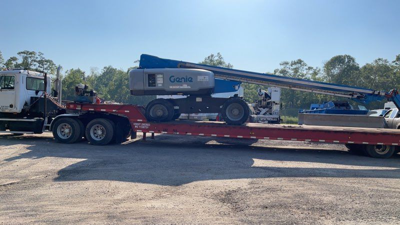 Blue boom lift on a flatbed trailer at a construction site, being hauled by a truck.