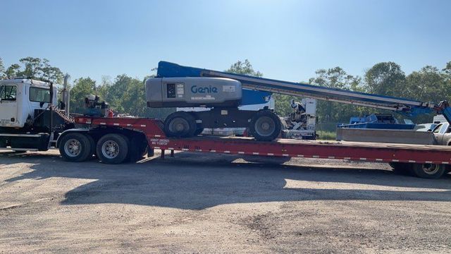 Blue boom lift loaded on a red flatbed trailer beside a white semi-truck in a gravel lot