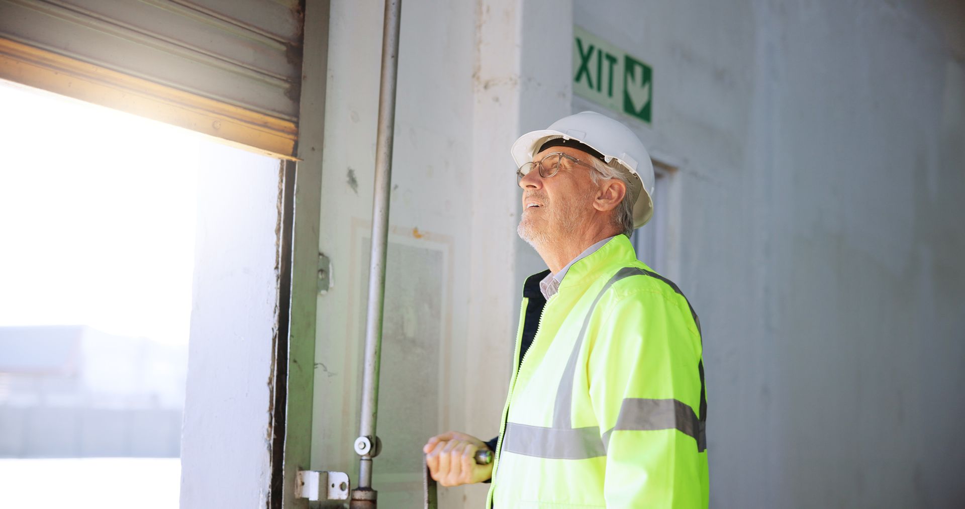 Worker in hard hat and reflective jacket inspecting an industrial doorway beneath an exit sign.