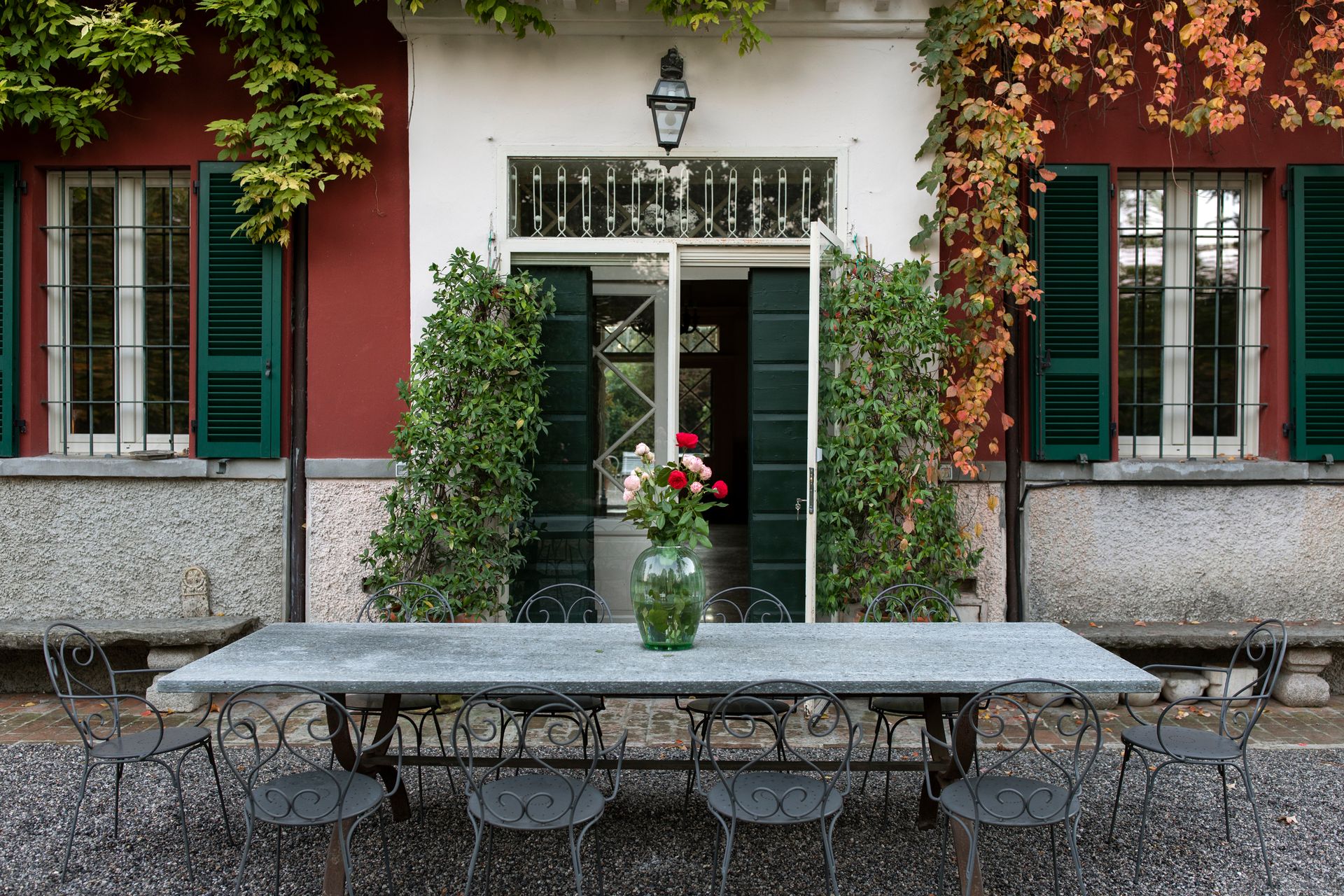 Red stucco building with green shutters, vine-covered entrance, outdoor dining table with flowers.