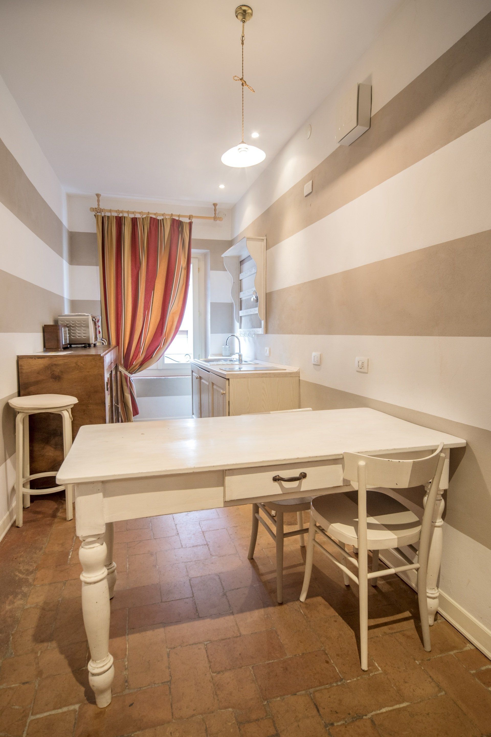 Tavolo da pranzo bianco con sedia in una stanza con pareti a strisce, tende e una piccola zona cucina.