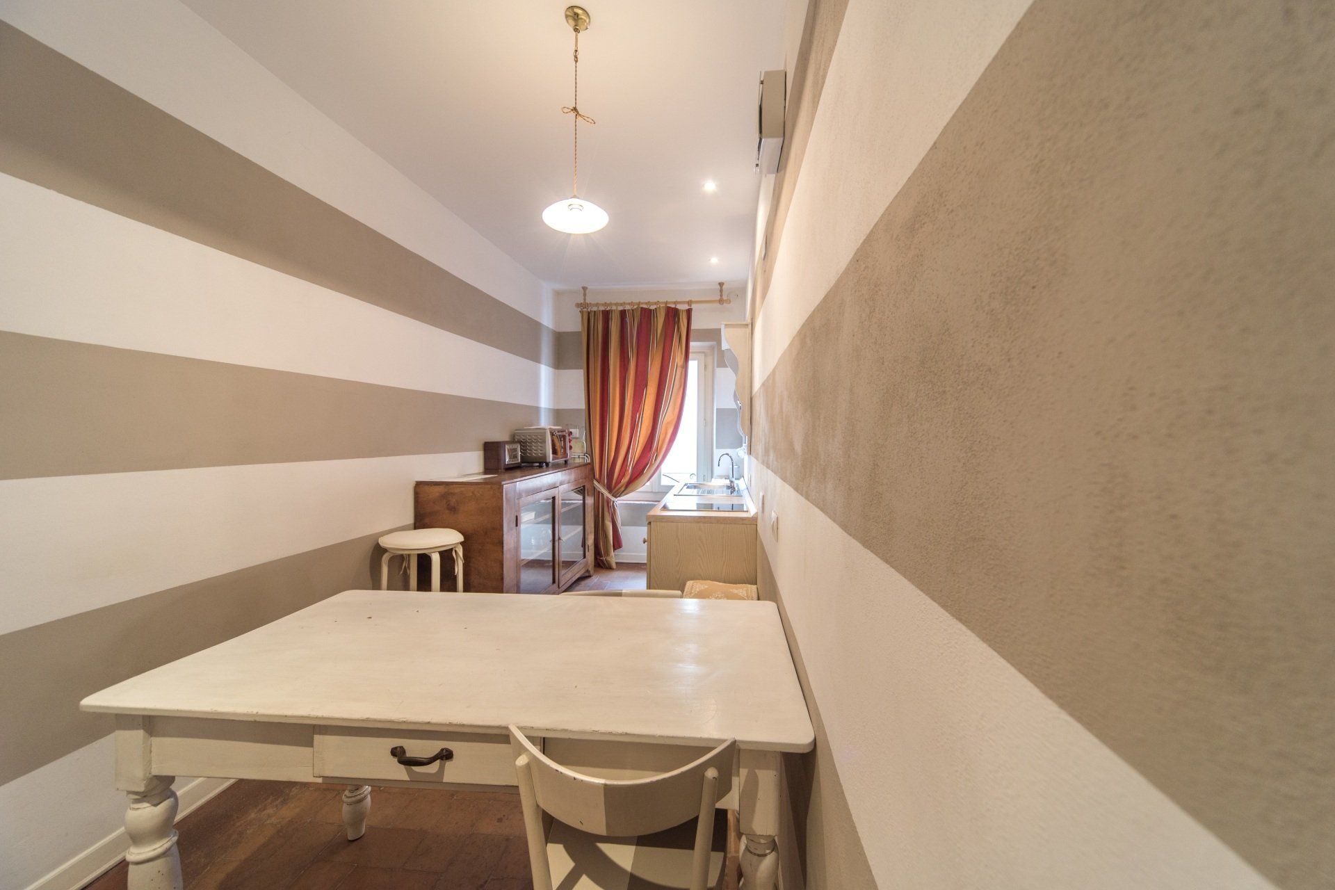 Piccola cucina con pareti a strisce bianche e beige, un tavolo bianco e un mobiletto in legno.