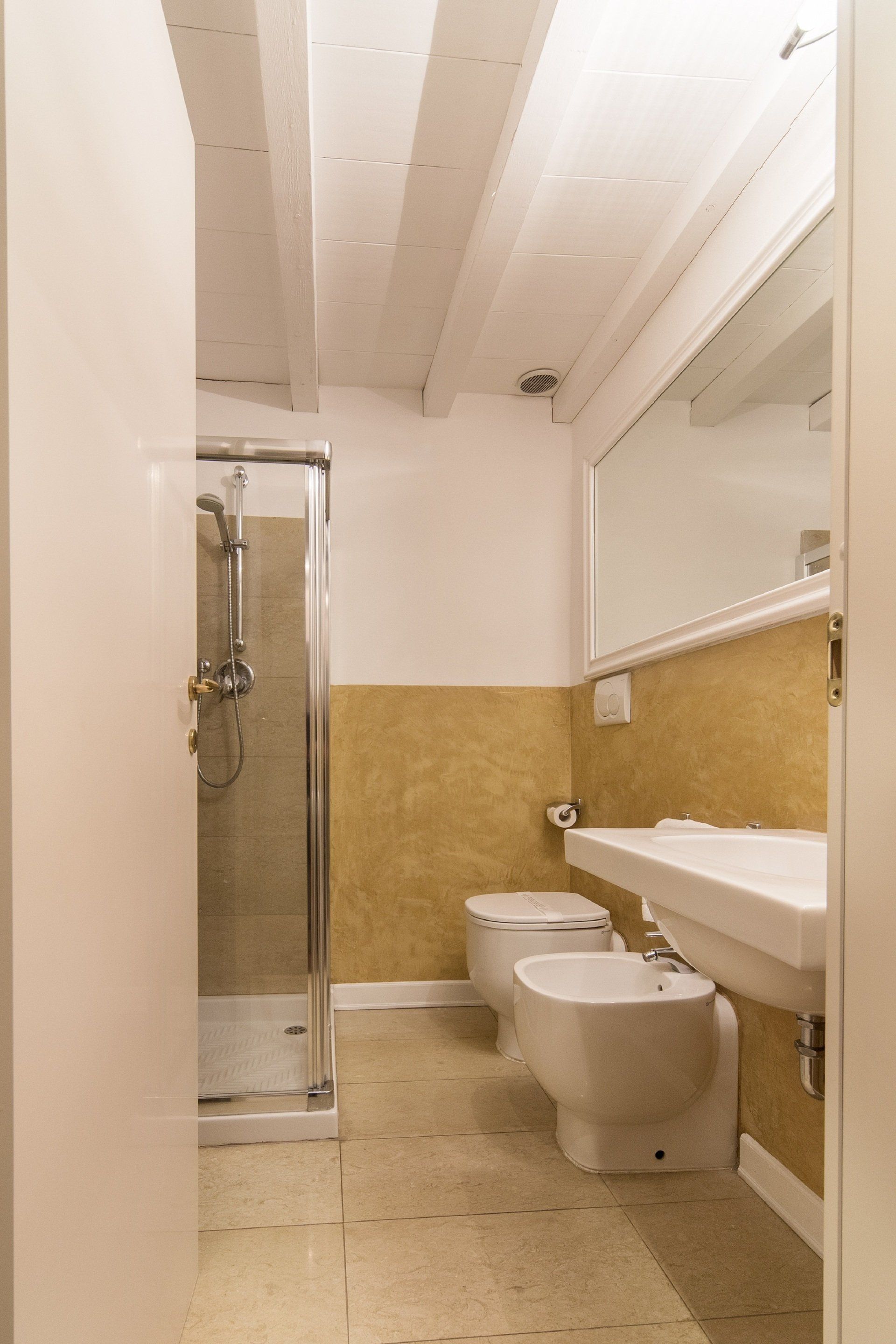 Bagno con doccia, WC e lavandino. Parete gialla, travi bianche al soffitto e pavimento beige.