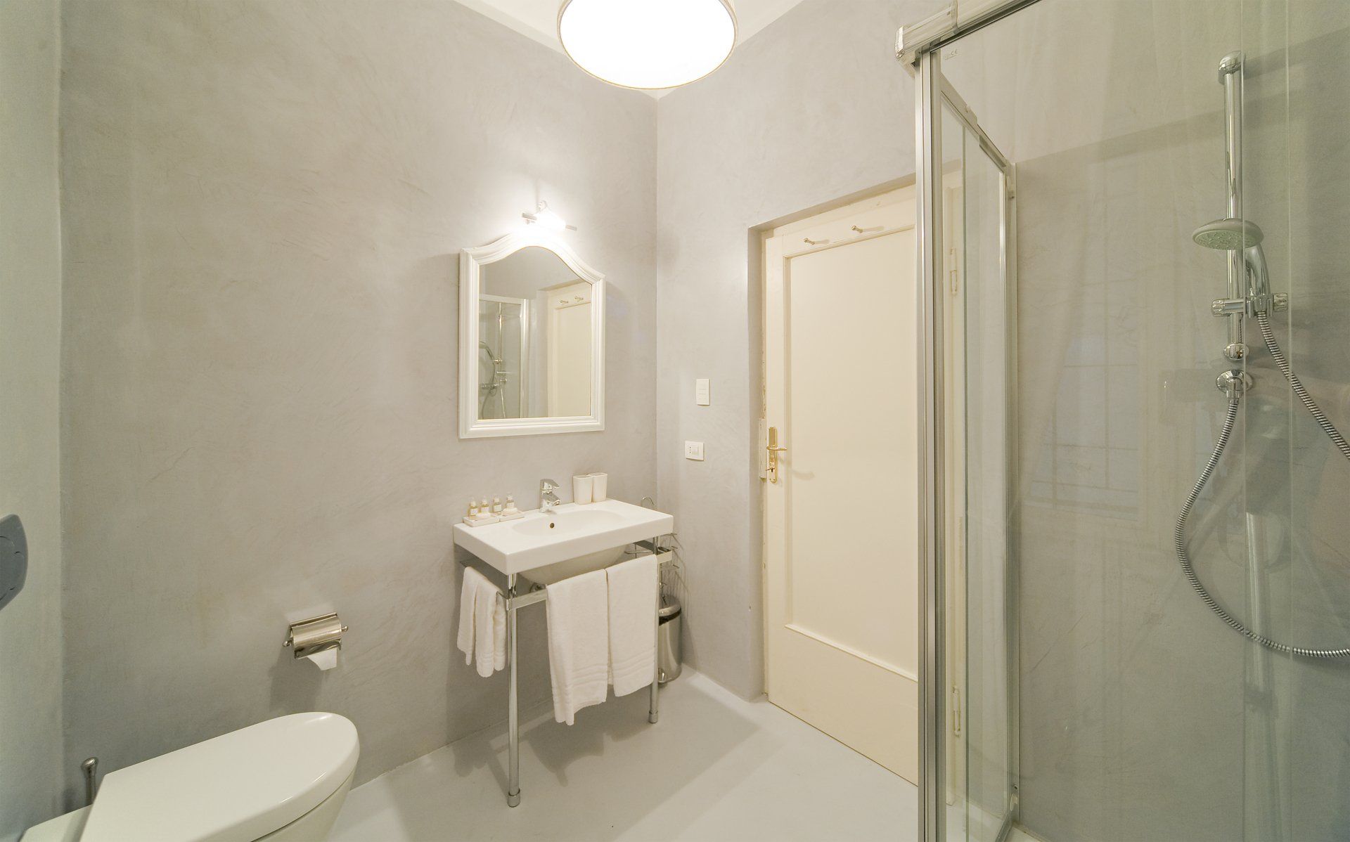 Bagno con wc, lavandino, doccia bianchi e pareti grigio chiaro. Si vede una porta bianca.