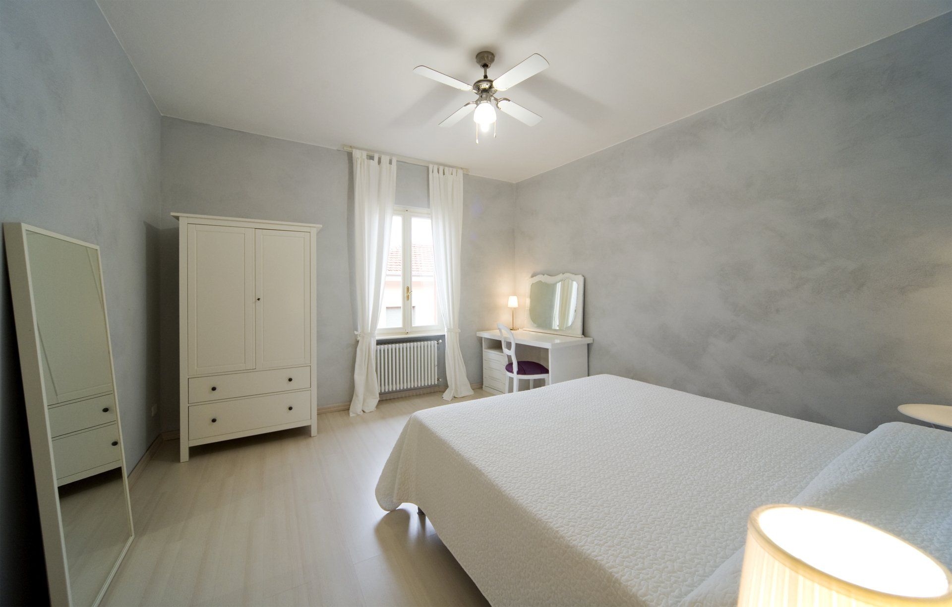 Camera da letto con letto bianco, armadio e mobiletto per toeletta su pareti grigie; finestra con tende.