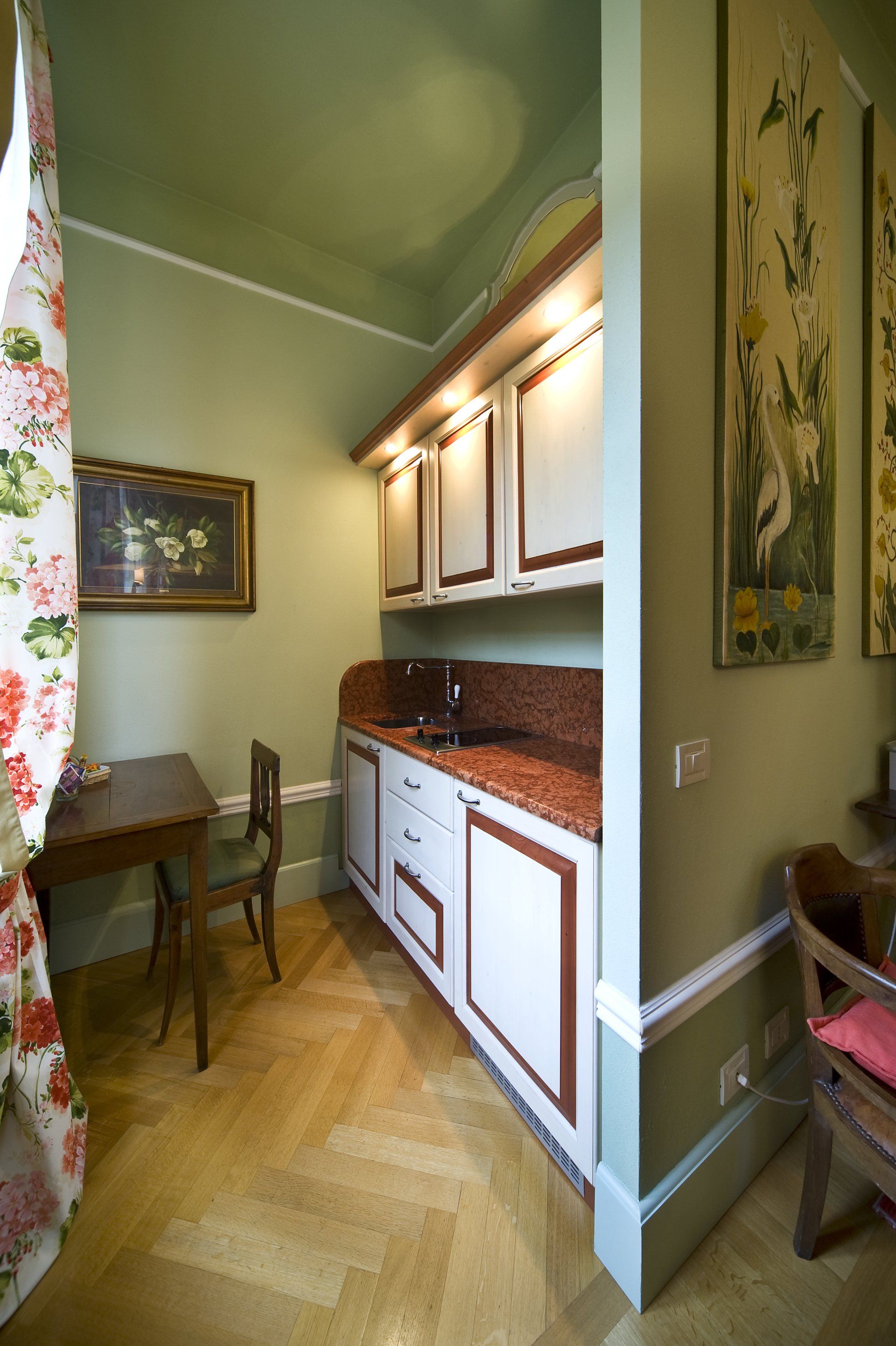 Piccola cucina con mobili bianchi, piano di lavoro in granito, pavimenti in legno, pareti verdi e opere d'arte.