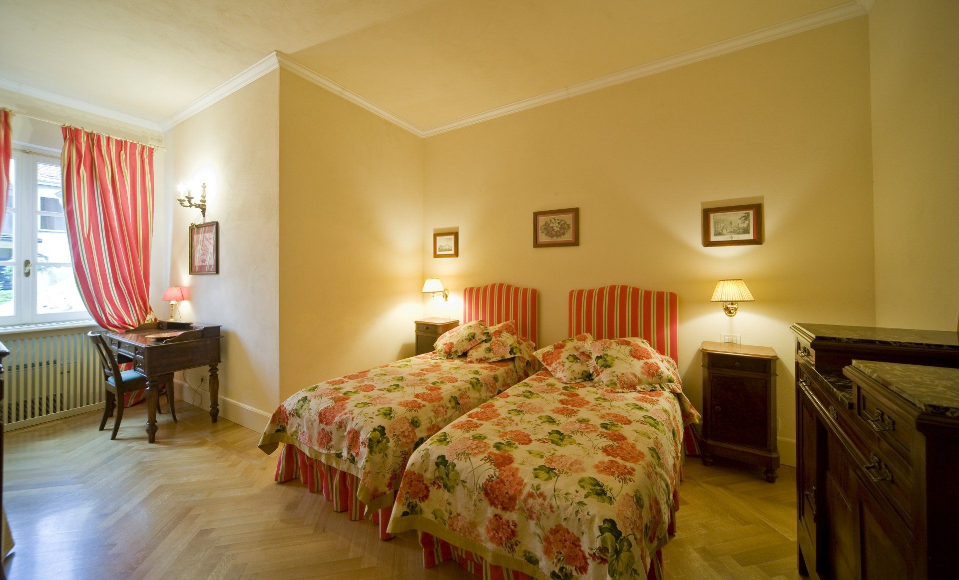 Una camera da letto con due letti singoli, biancheria da letto floreale, mobili in legno e tende a righe rosse e bianche.