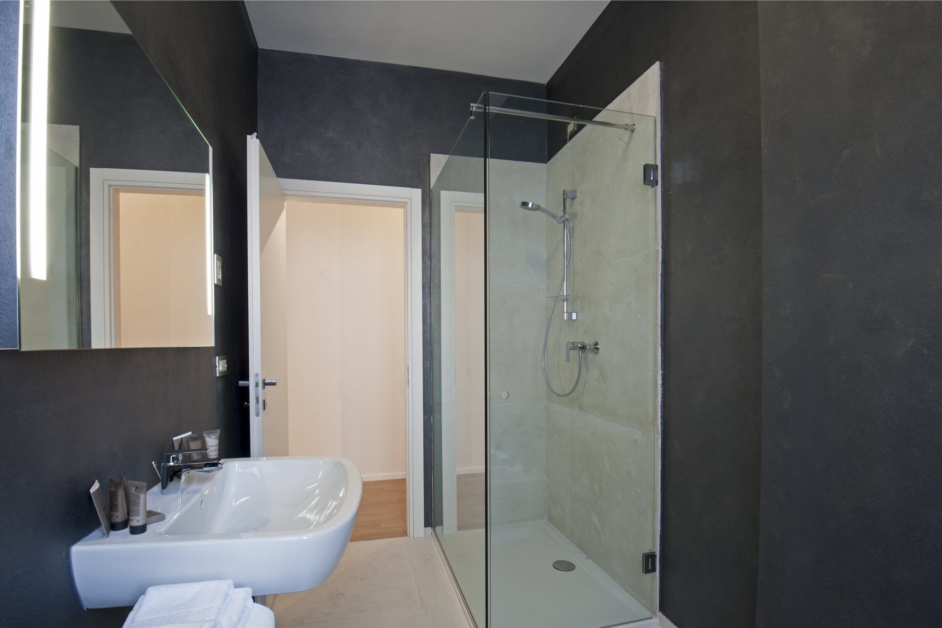 Bagno con pareti grigie, doccia in vetro, lavandino bianco e porta aperta su un'altra stanza.