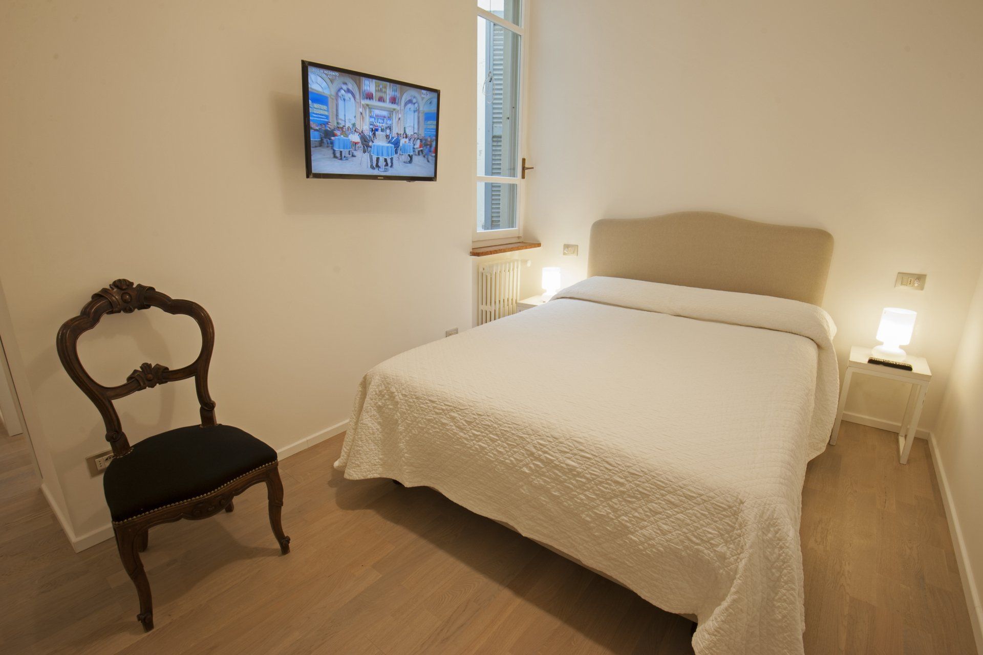 Camera da letto con letto matrimoniale, TV, sedia antica e comodino. Pareti chiare, pavimento in legno.