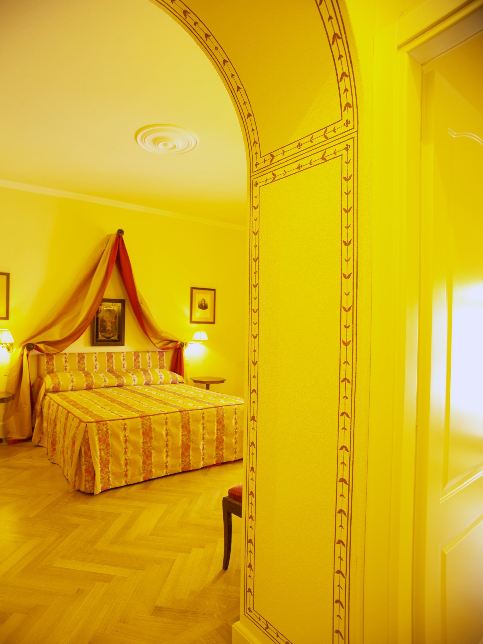 Interno di una camera da letto con un letto, attraversato da un arco. Toni gialli, baldacchino sopra il letto.