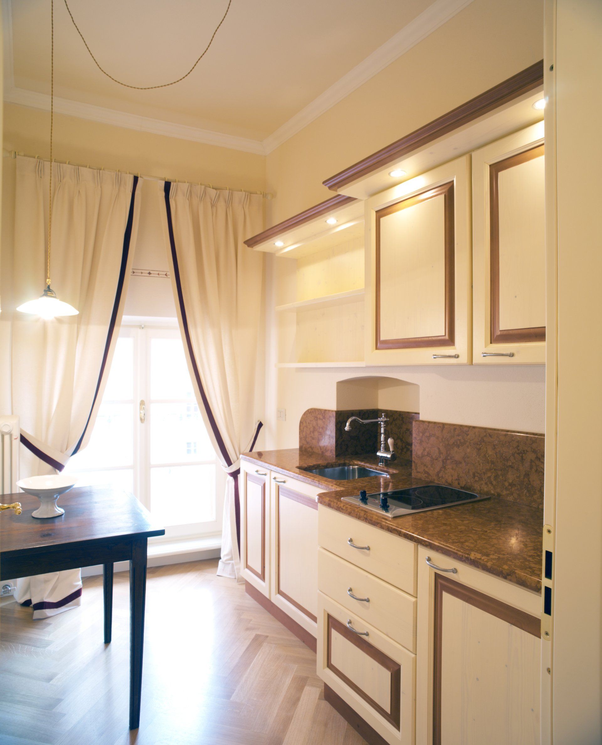 Cucina color crema con dettagli in legno, mobili e tavolo scuro. Finestra con tende bianche.