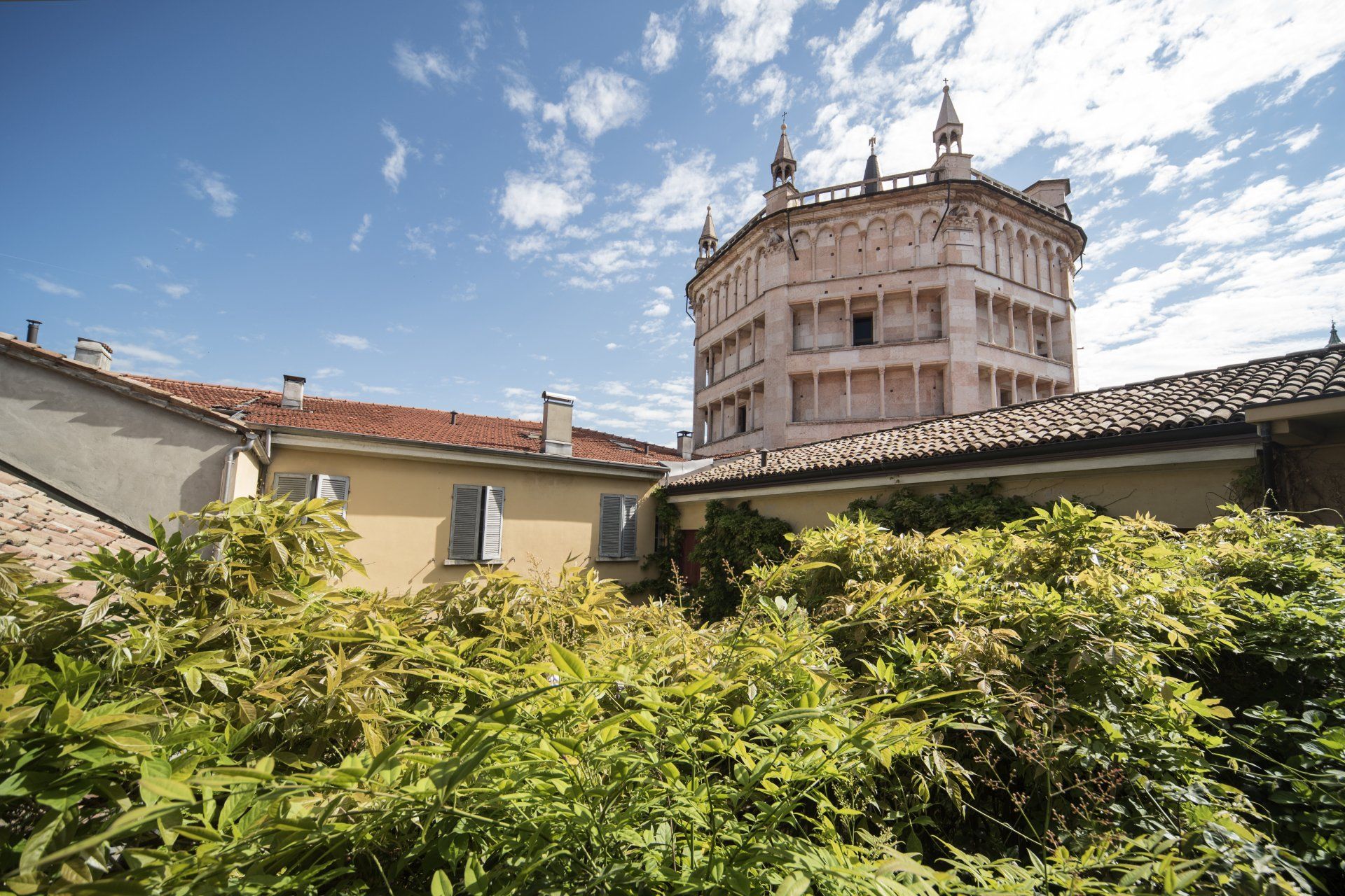 Il Battistero ottagonale di Parma dietro fitti cespugli verdi e un edificio giallo sotto il cielo azzurro.