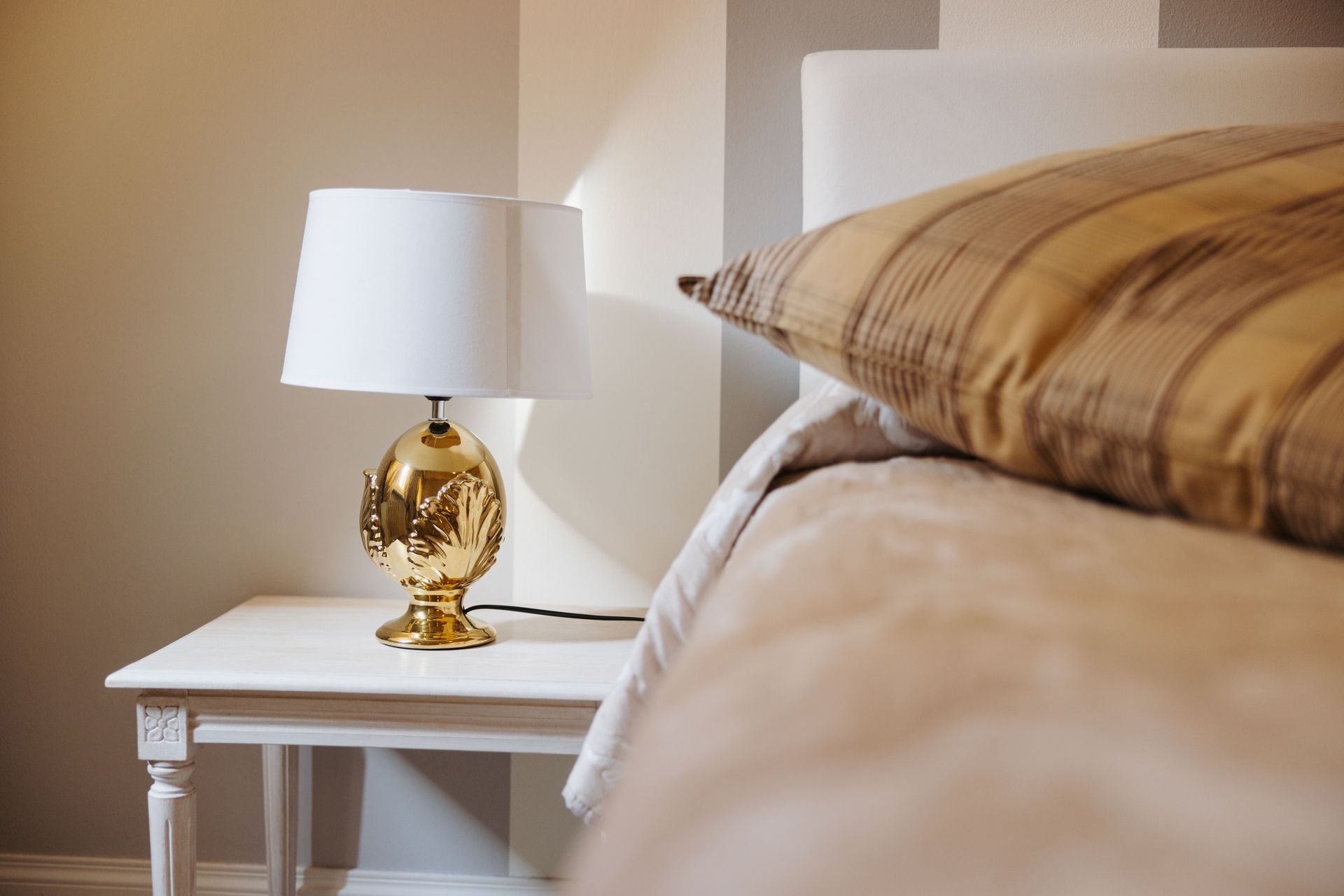 Lampada a forma di ananas dorato sul comodino bianco accanto al letto con cuscino color cuoio.