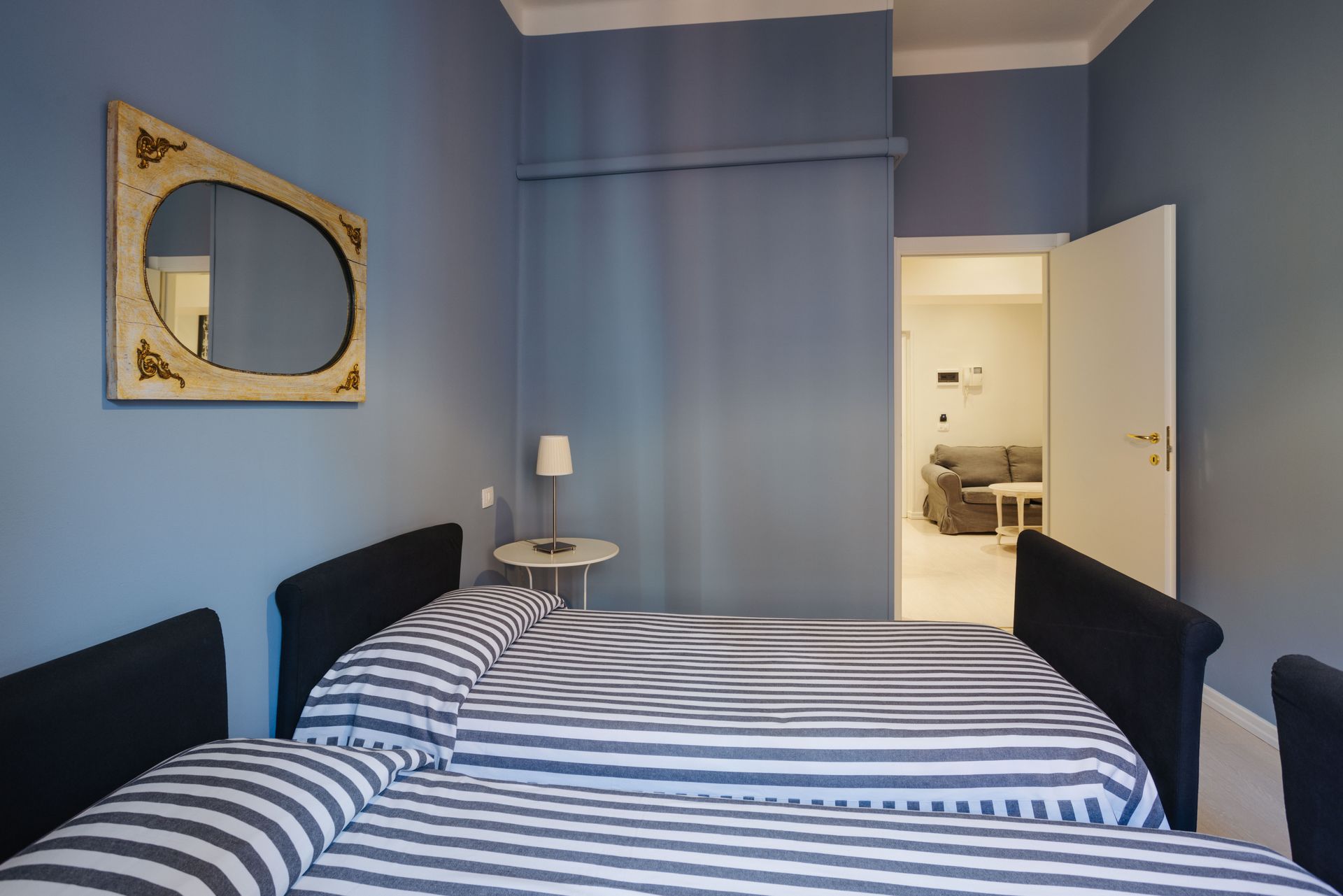 Camera da letto con pareti blu, un letto a righe, una porta aperta su un'altra stanza e uno specchio decorativo.