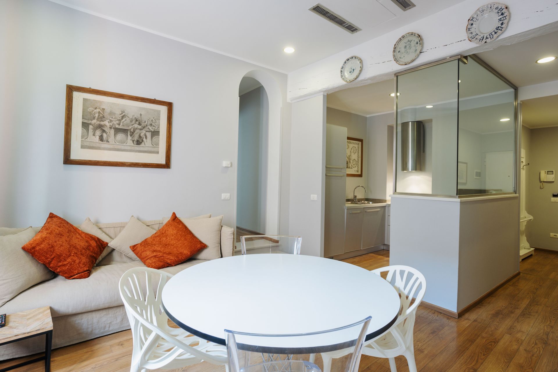 Soggiorno con divano, tavolo rotondo, cucina dietro una vetrata. Arredamento beige, arancione e bianco.