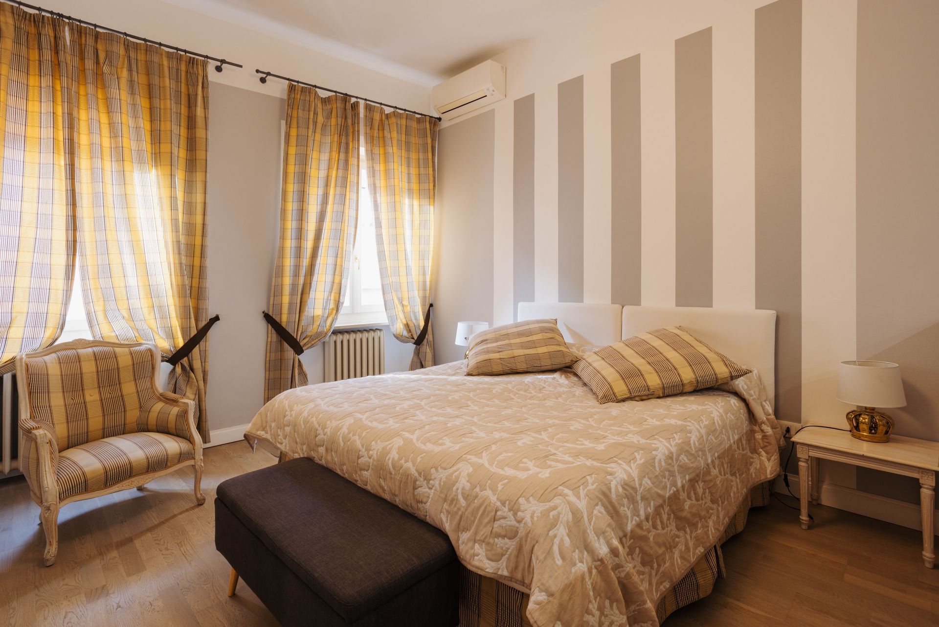 Camera da letto con parete a strisce, letto con biancheria a fantasia, sedia, tende e tavolino con lampada.