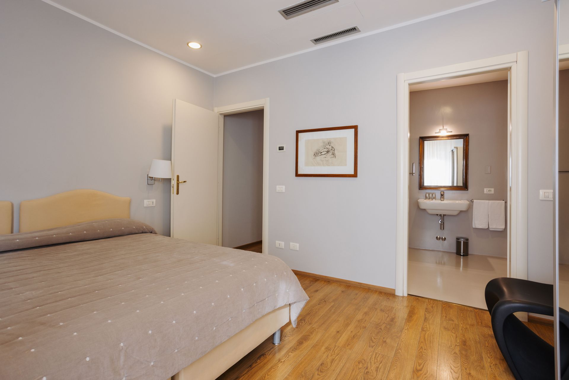 Camera da letto con un letto, due porte e un bagno. Pareti grigio chiaro, pavimento in legno.