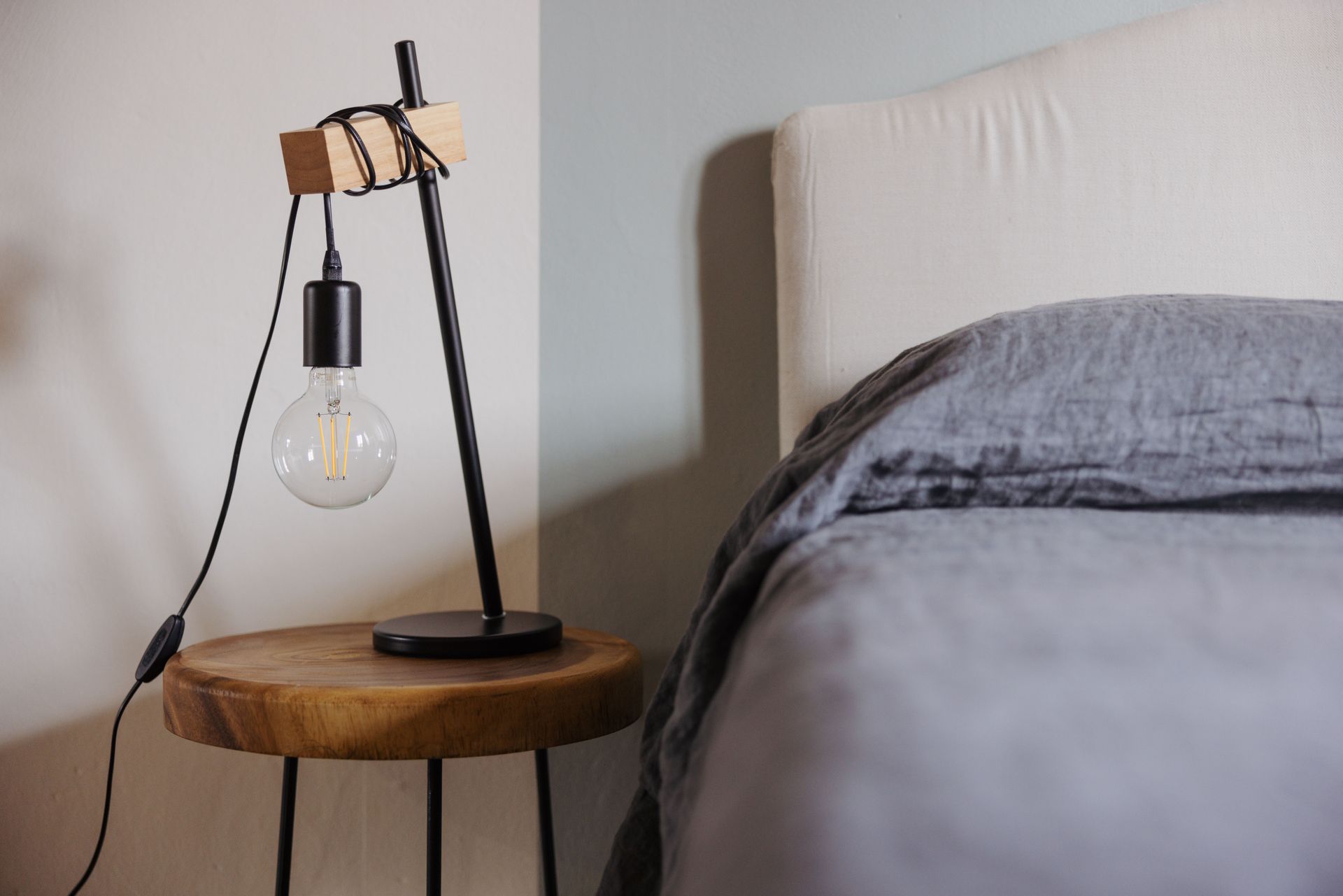 Comodino con una lampada nera unica e un letto con lenzuola grigie.