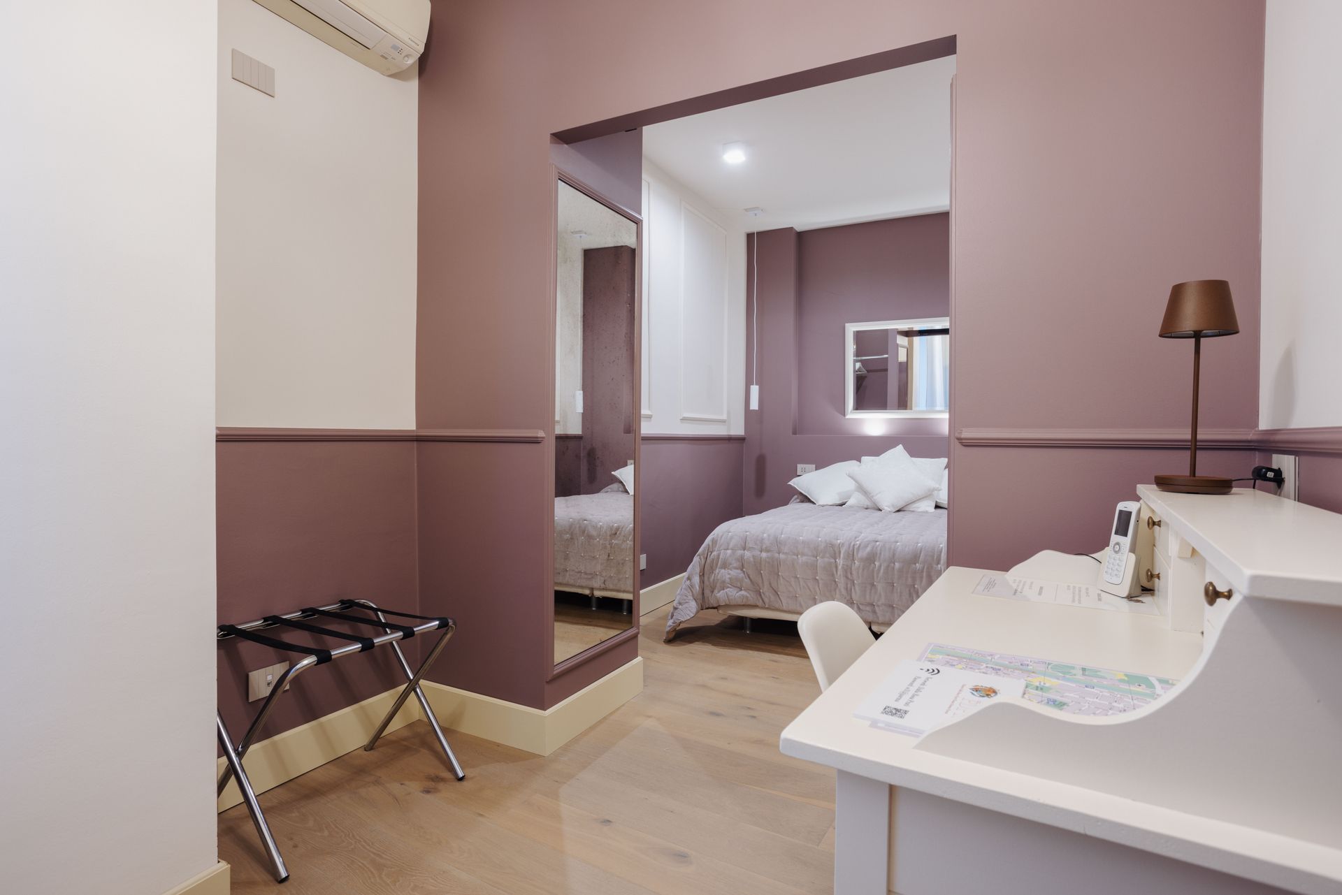 Interno di una camera d'albergo con scrivania, portavaligie e un letto visibile attraverso una porta.
