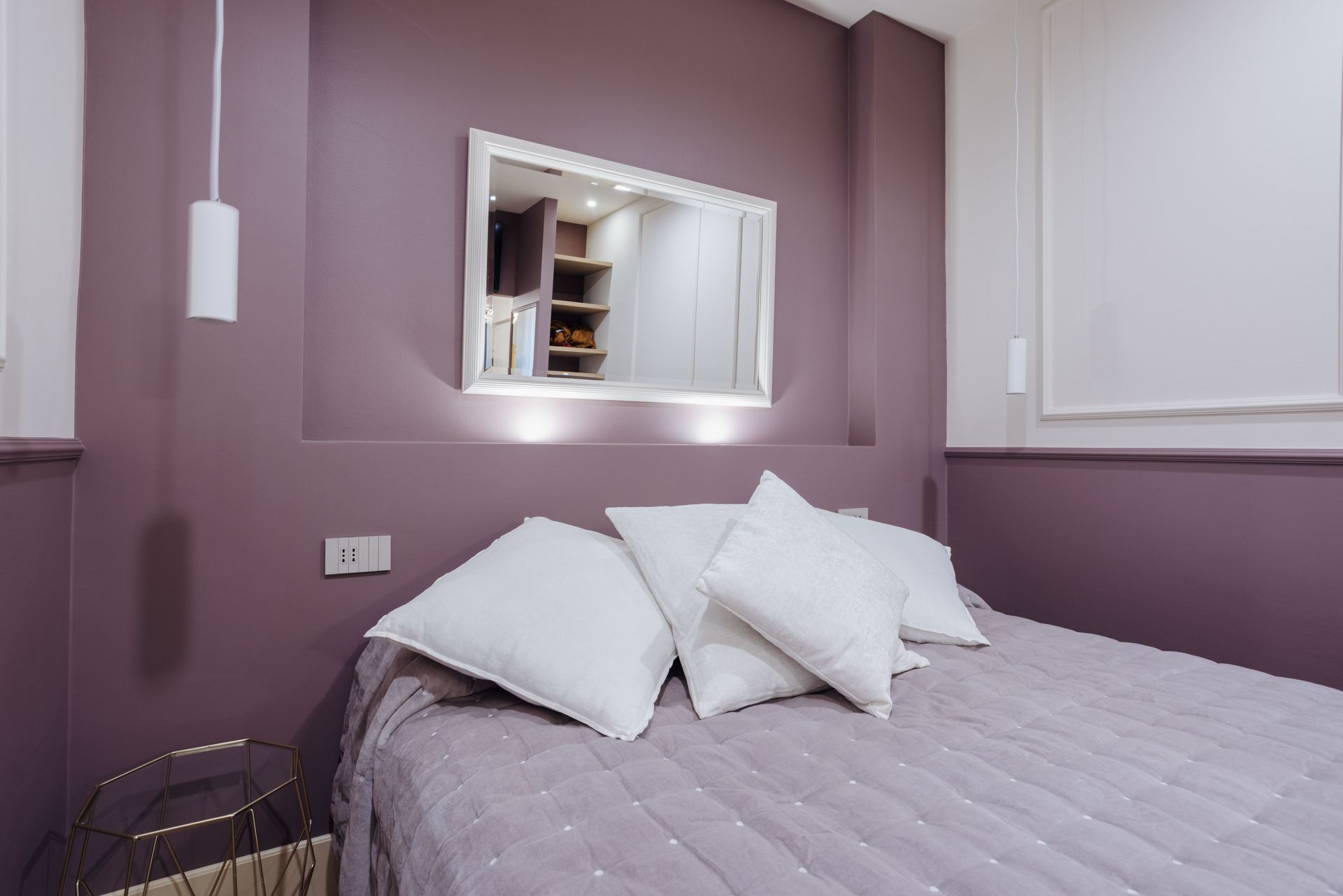 Camera da letto con pareti color lavanda, biancheria da letto e cuscini bianchi, specchio e comodino.