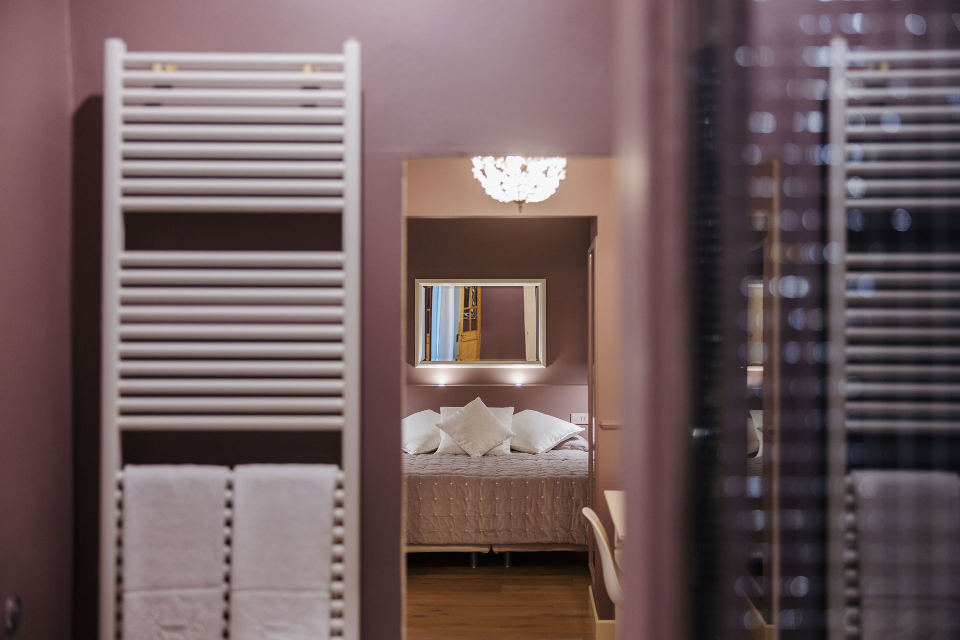 Bagno con portasciugamani bianco e vista sulla camera da letto con letto, specchio e lampadario.
