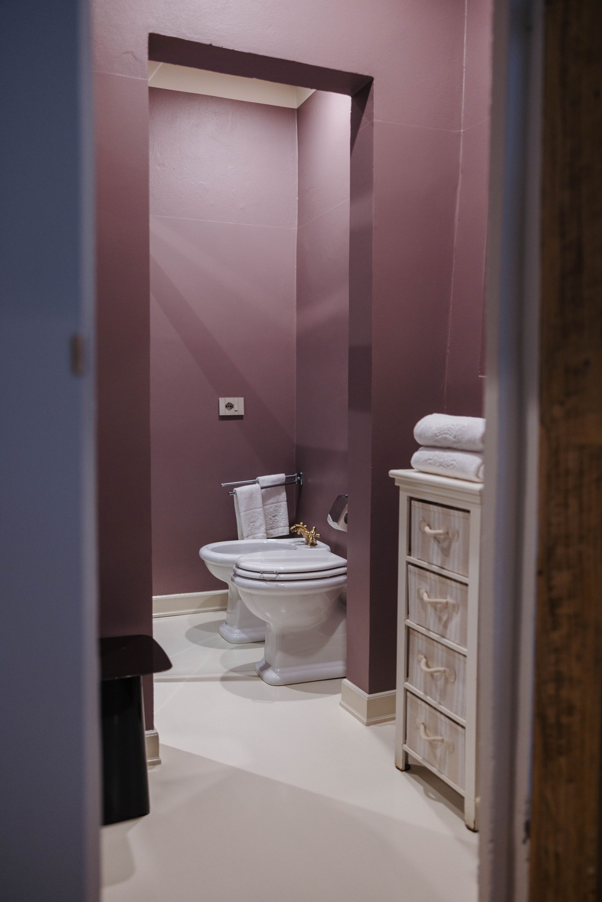 Un piccolo bagno dipinto di malva con wc bianco, bidet e mobiletto portaoggetti.