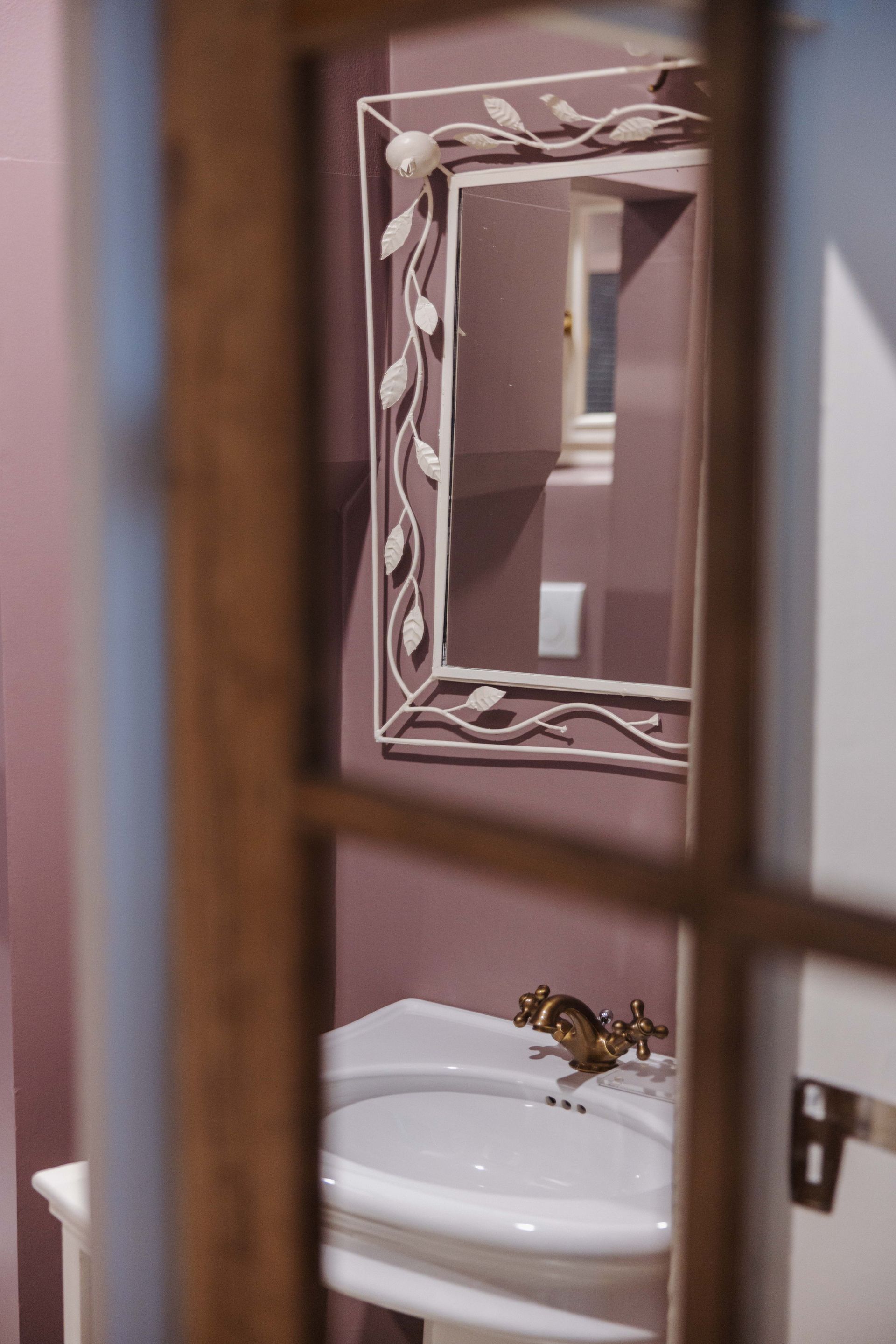 Un piccolo bagno, visto attraverso una cornice di legno, con lavandino, specchio e pareti color malva.