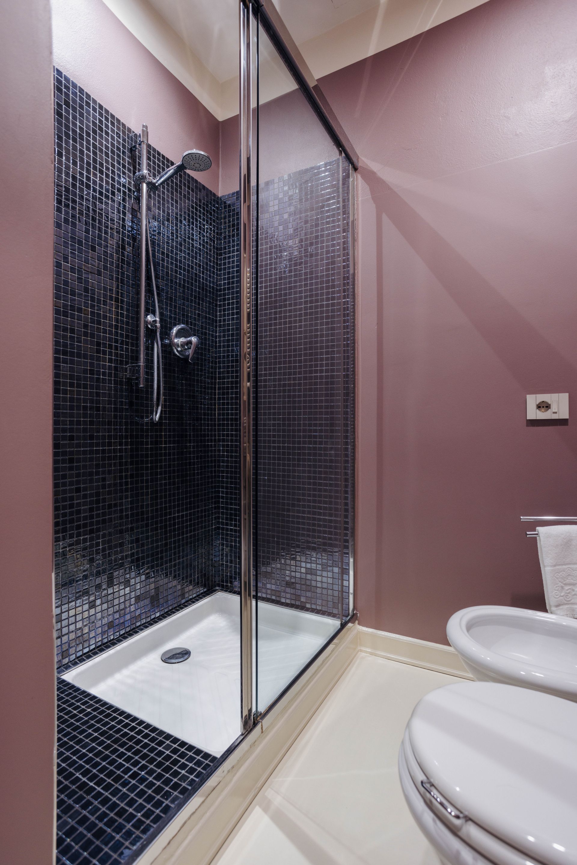 Doccia con piastrelle nere e porta in vetro, accanto a bidet e WC. Pareti viola.