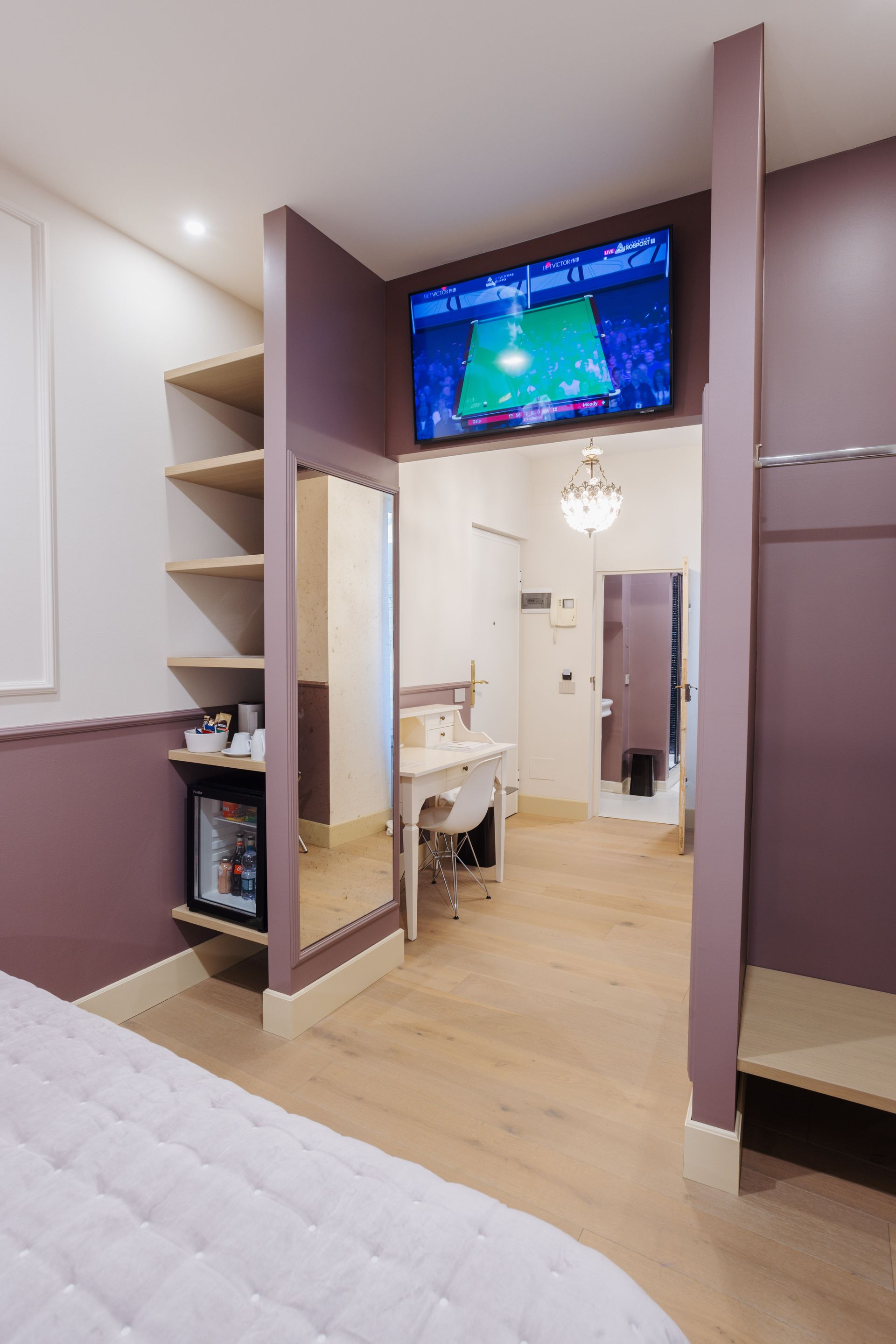 Interno di una camera d'albergo con TV, scaffali aperti, frigorifero e vista su una scrivania e un bagno.