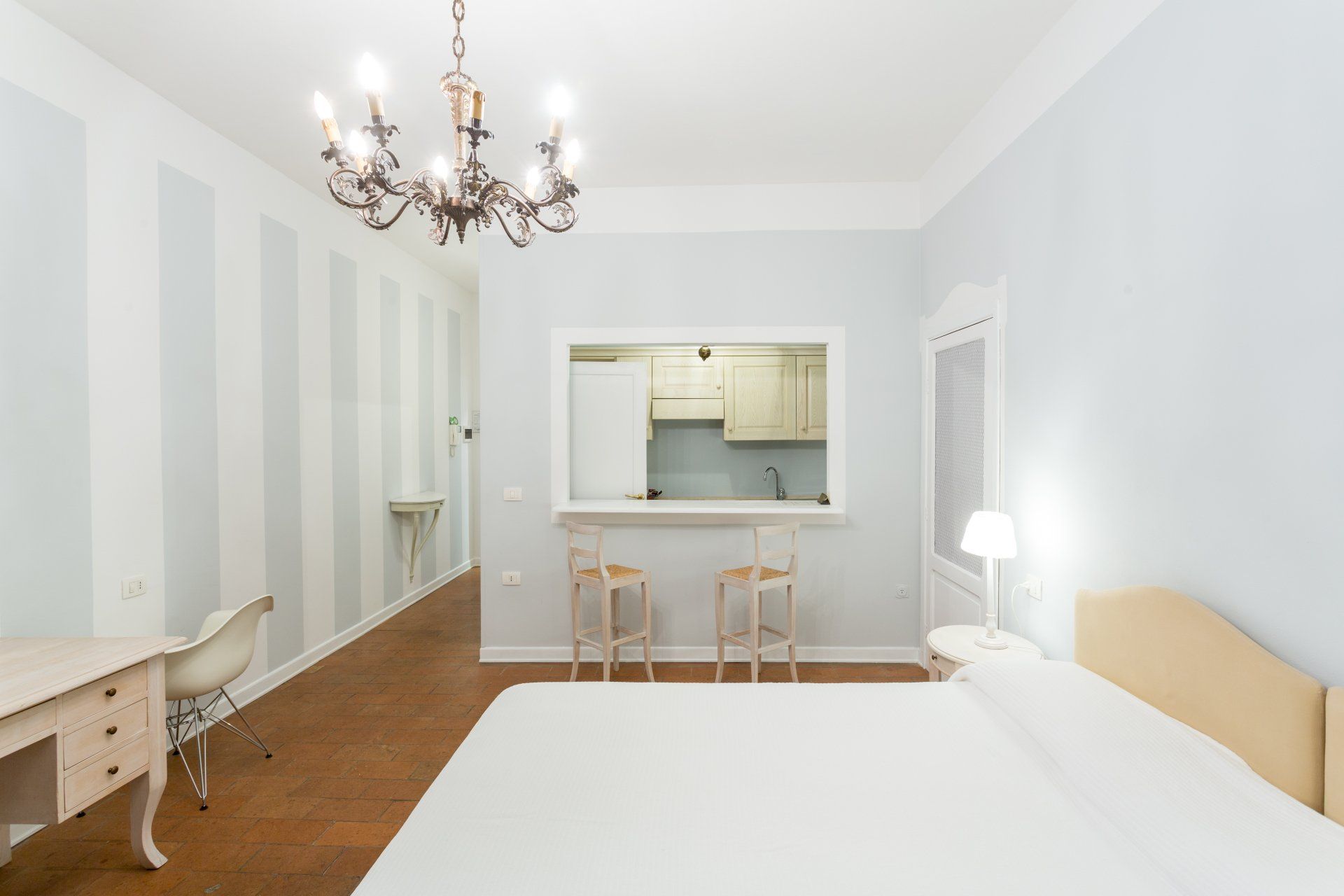 Camera da letto con letto bianco, scrivania e passaggio per la cucina. Parete a strisce, lampadario.