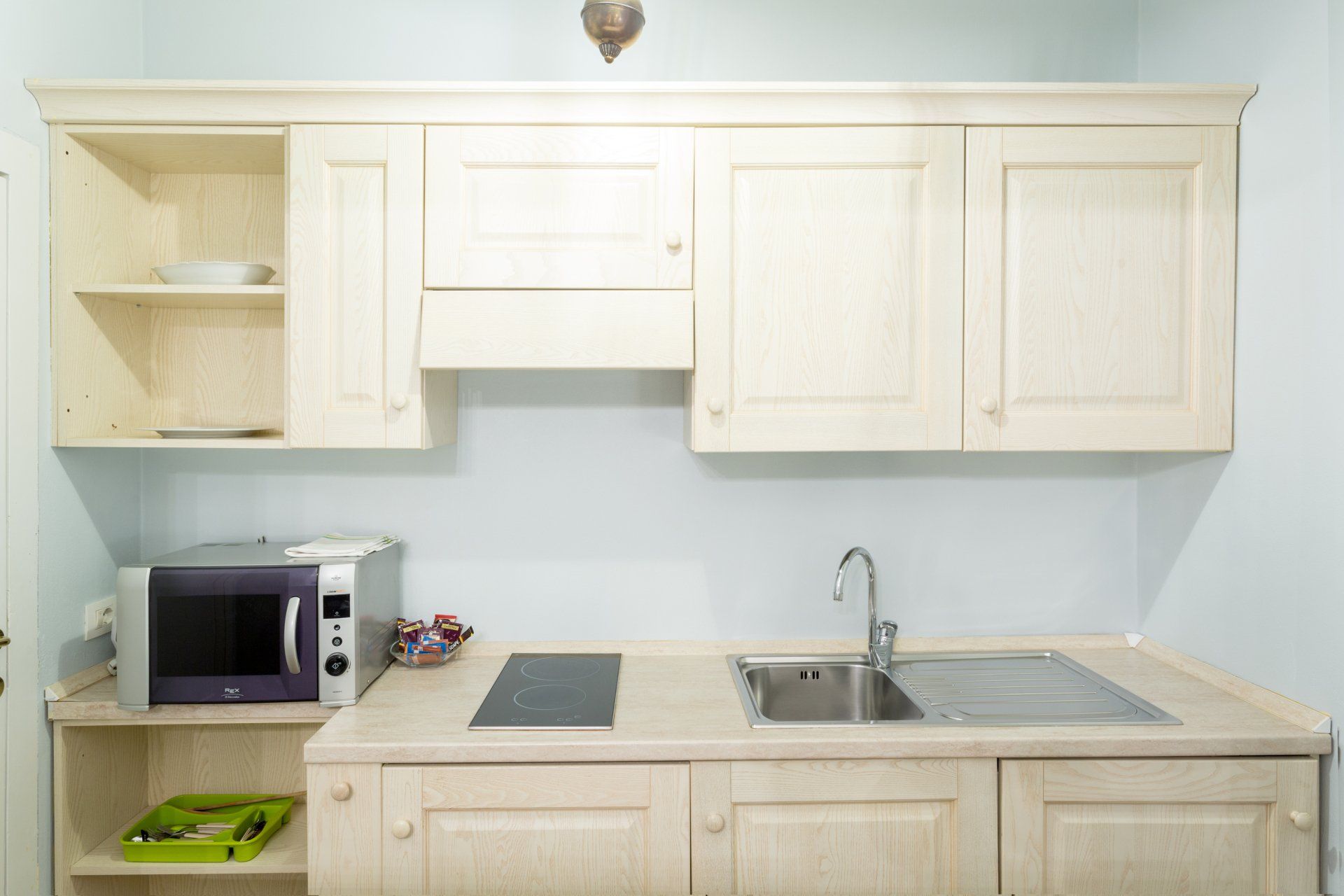Piccola cucina con mobili in legno chiaro, lavello, microonde e piano cottura.