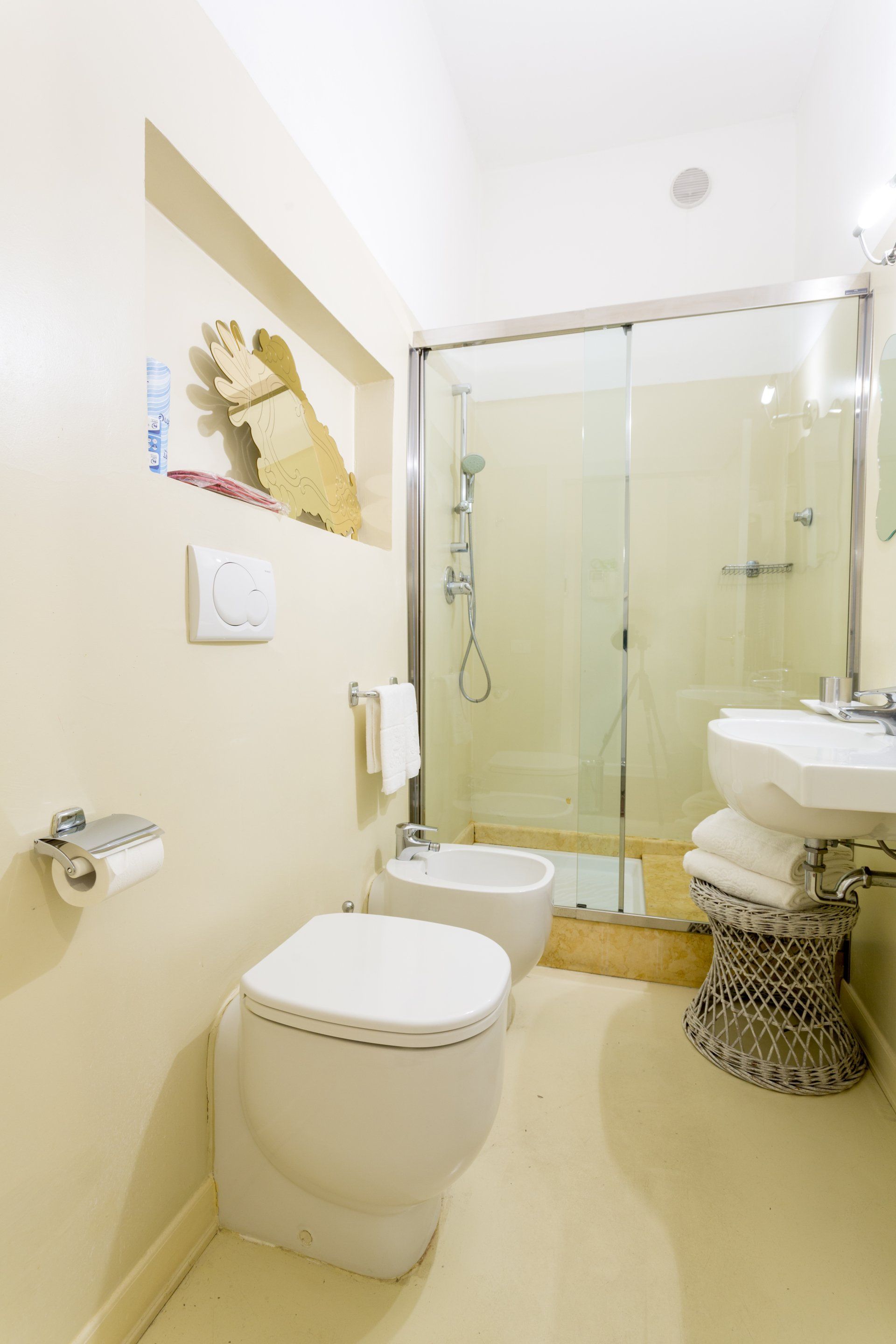 Bagno color crema con WC, bidet e doccia. Un lavandino con asciugamani su uno sgabello decorativo.