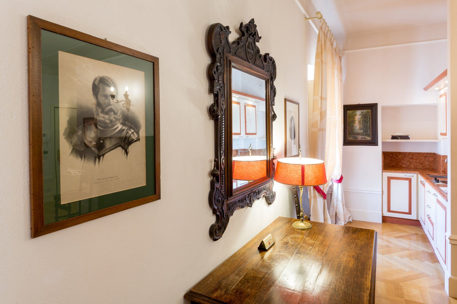 Corridoio con quadri incorniciati, uno specchio decorato e una lampada su un tavolo di legno, che conduce alla cucina.