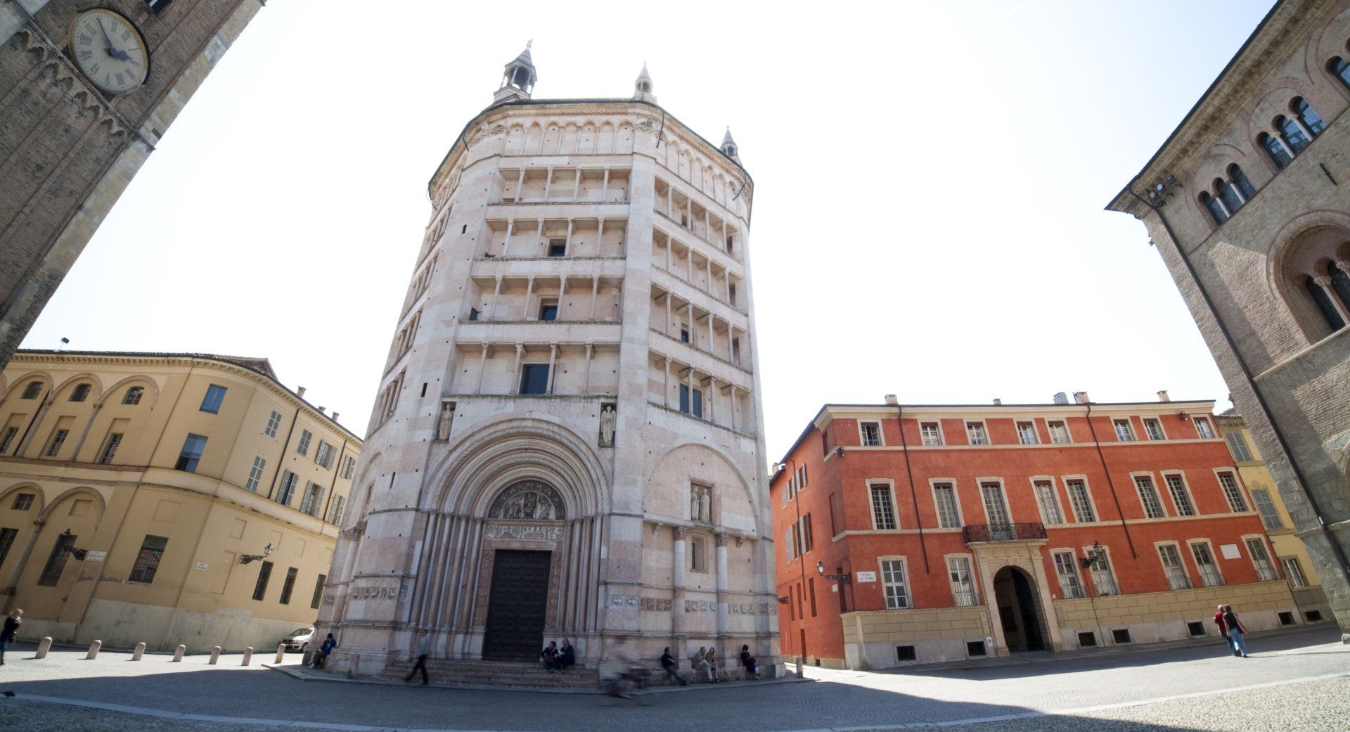 Battistero ottagonale bianco a Parma, Italia, fiancheggiato da edifici sotto un cielo luminoso.