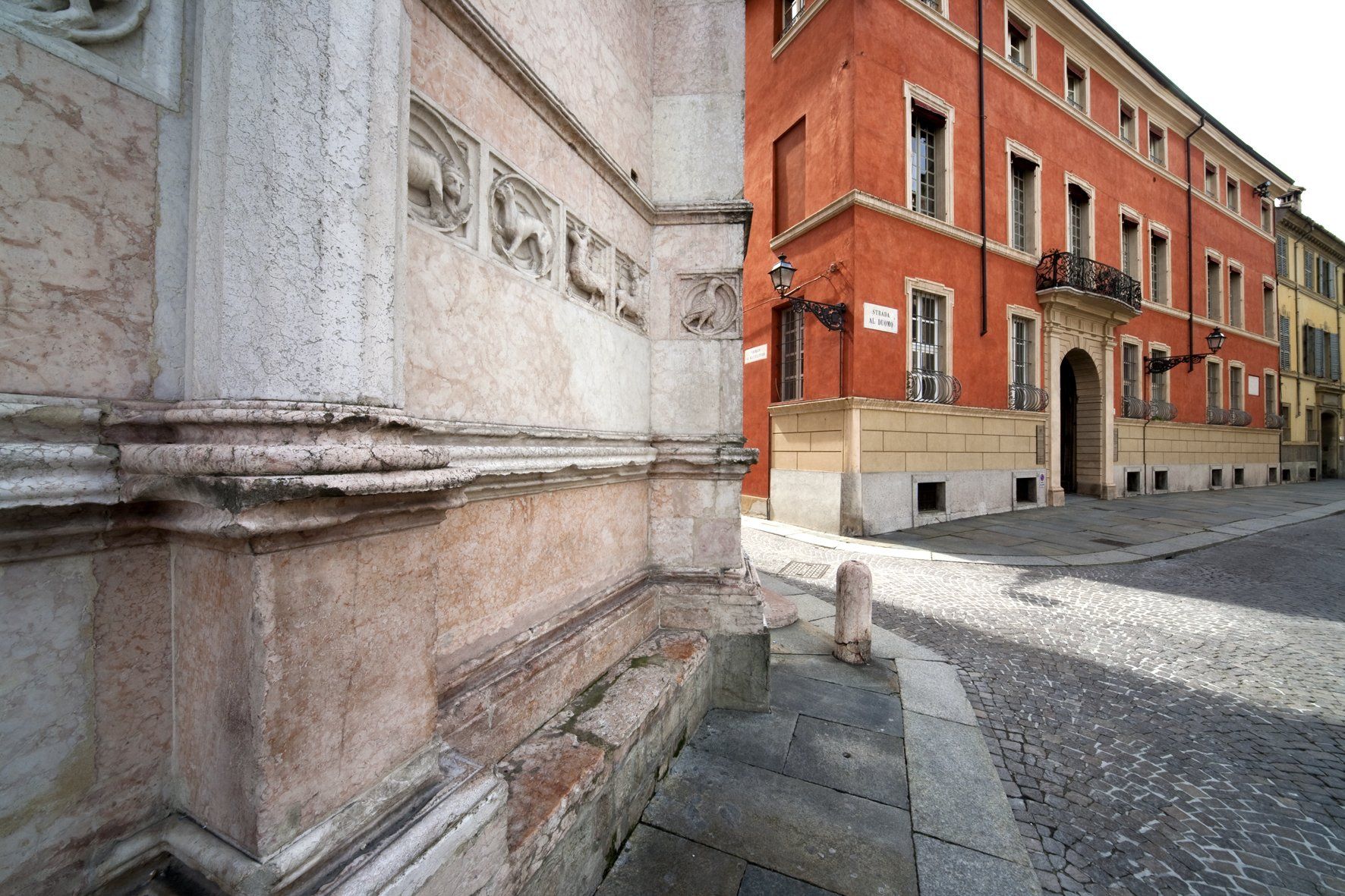 Angolo di un edificio decorato in pietra chiara con un edificio rosso e una strada acciottolata sullo sfondo.