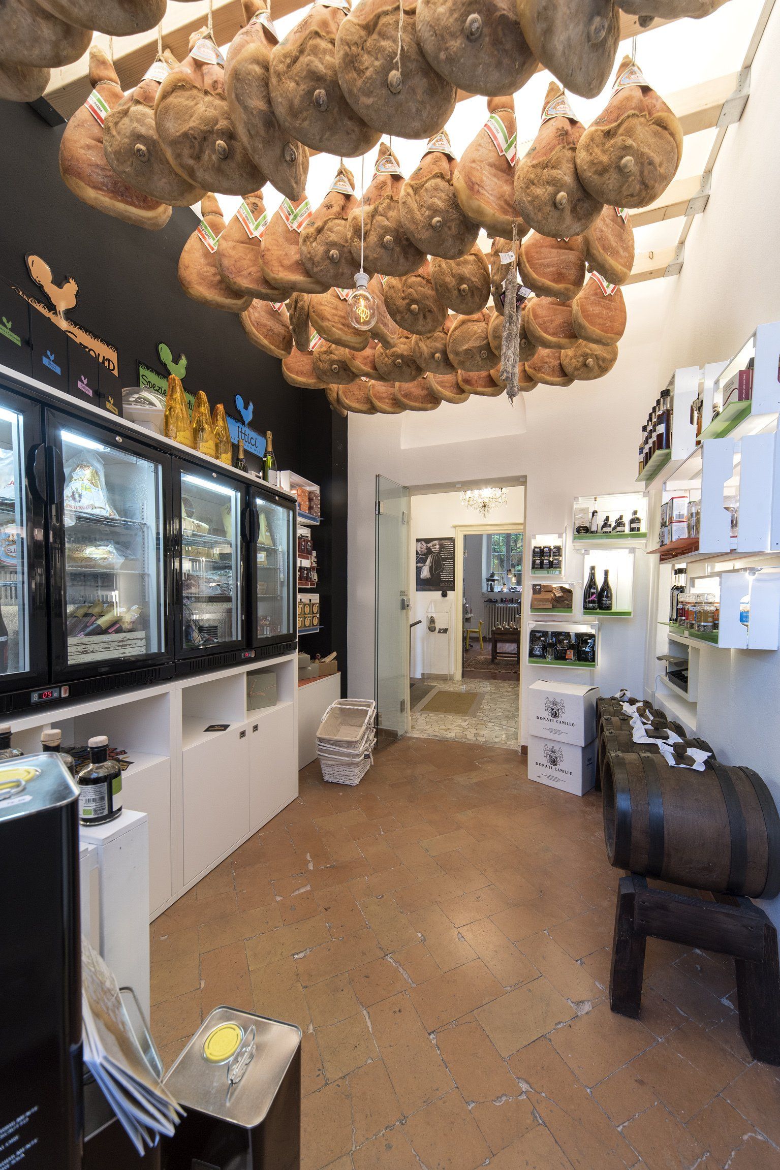 Interno di una gastronomia con carni appese, vetrine e una piccola selezione di vini; colori marrone e bianco.