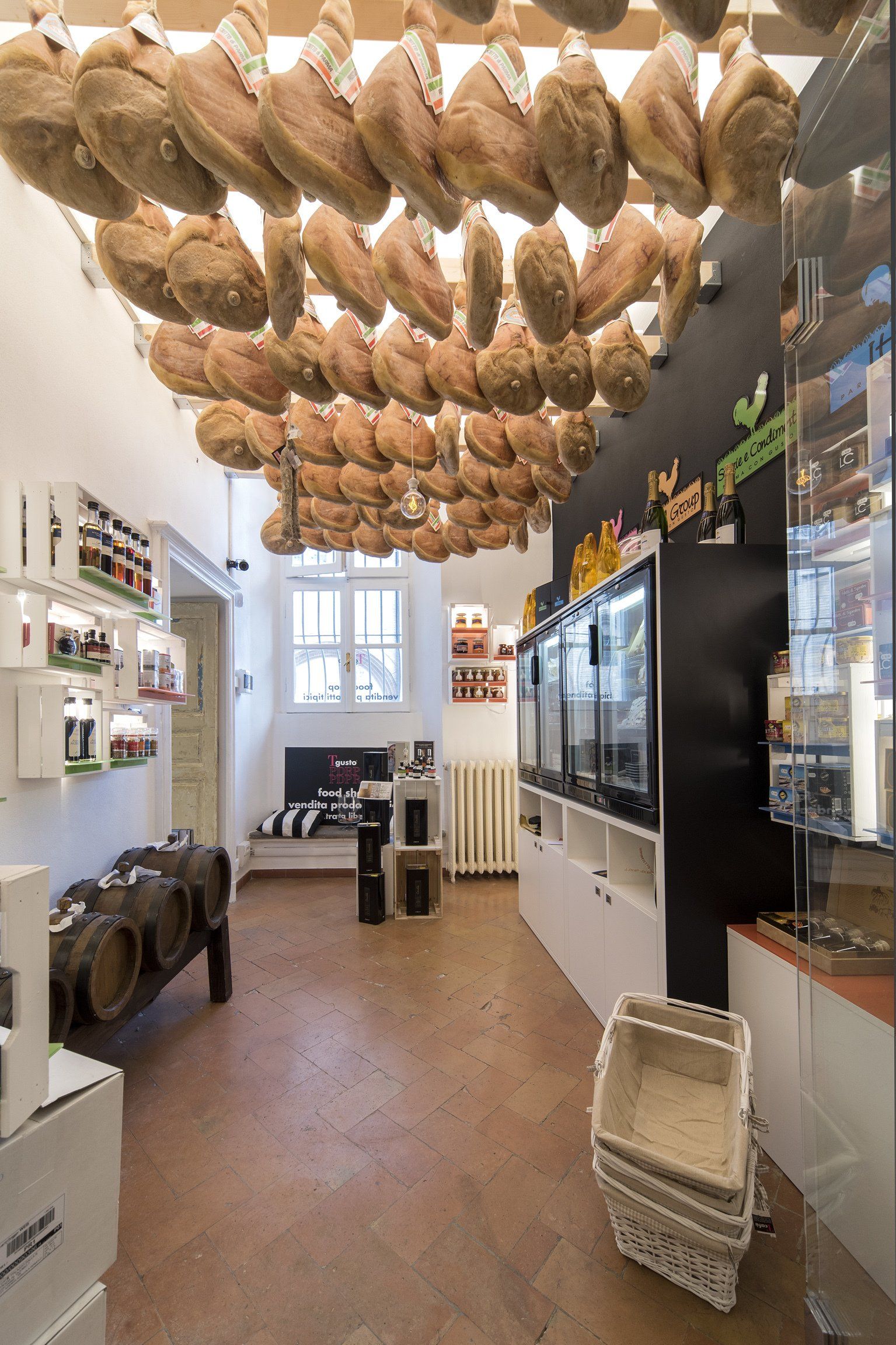 Interno di un negozio con salumi appesi al soffitto, scaffali pieni di prodotti e un cesto bianco sul pavimento.