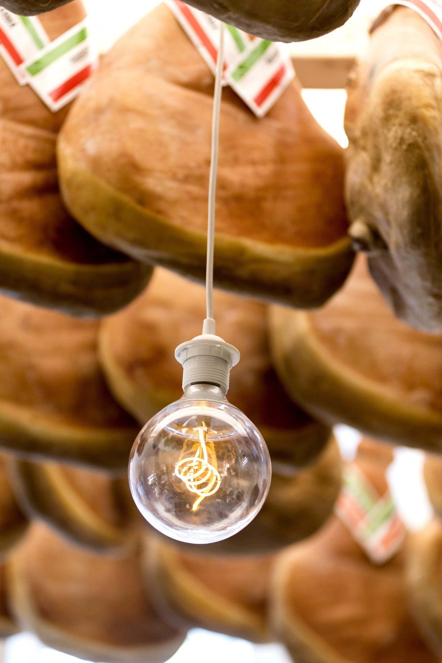 Lampadina appesa tra prosciutti stagionati appesi, con etichette raffiguranti la bandiera italiana, in un mercato.