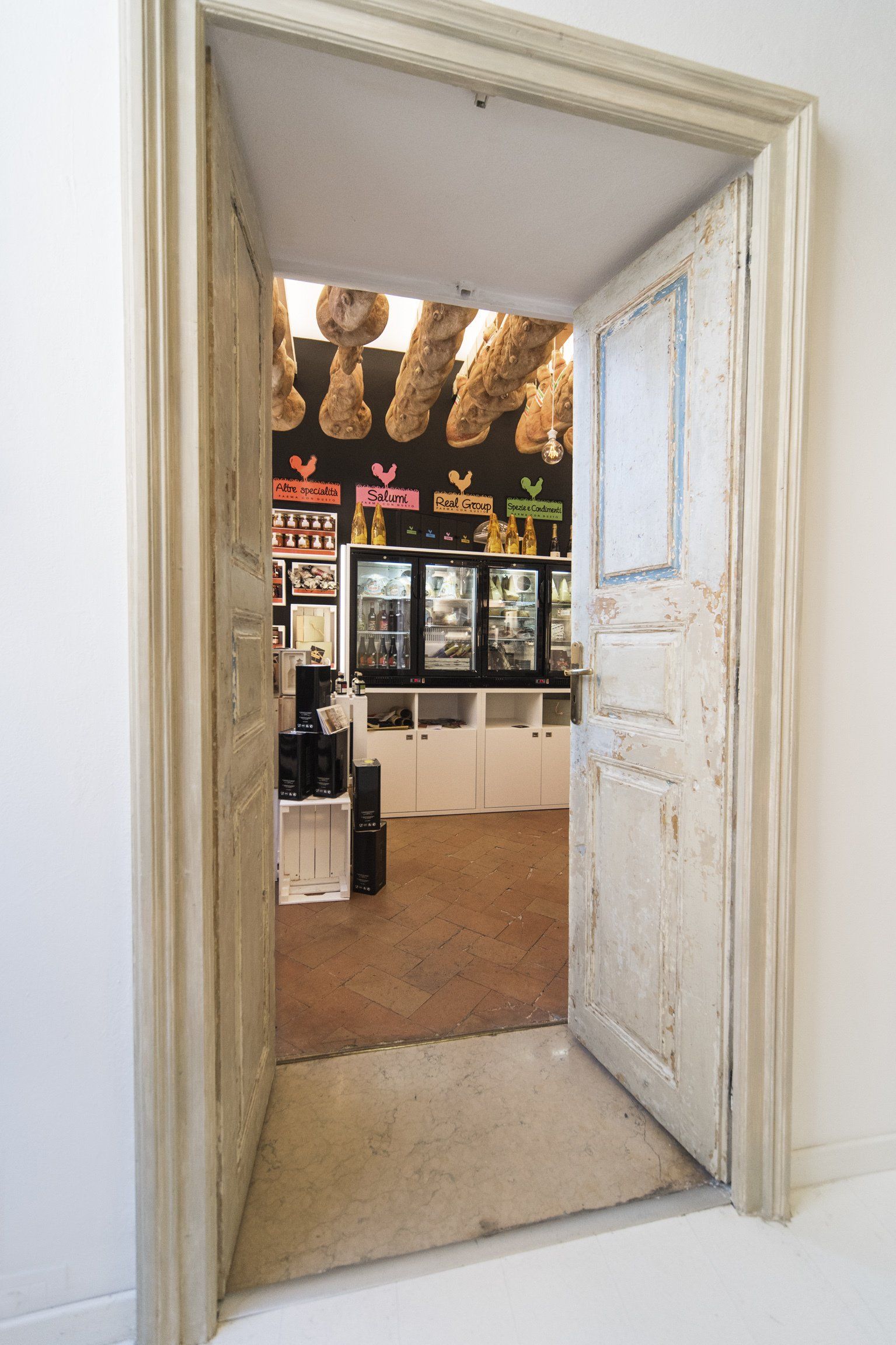 Una porta bianca si apre su un negozio; pavimento marrone, soffitto nero, merce esposta.