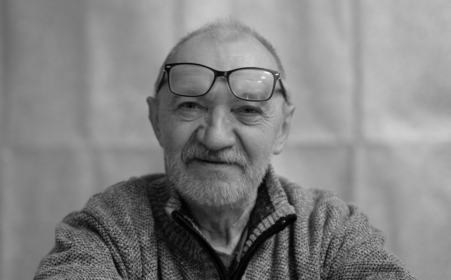 Smiling man with glasses resting on his head, wearing a textured sweater, against a plain backdrop
