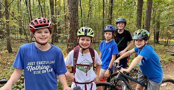 Five cyclists with helmets stand beside bikes on a forest trail, smiling at the camera.