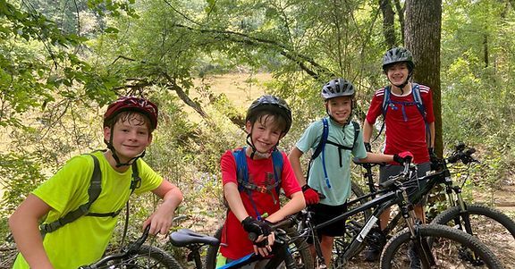 Four cyclists with helmets posing on mountain bikes in a sunny forest trail